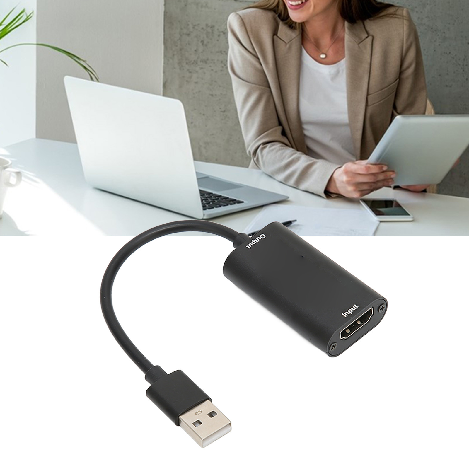 Ein schwarzer USB-zu-HDMI-Adapter im Vordergrund. Eine Frau benutzt einen Laptop und ein Tablet im Hintergrund.