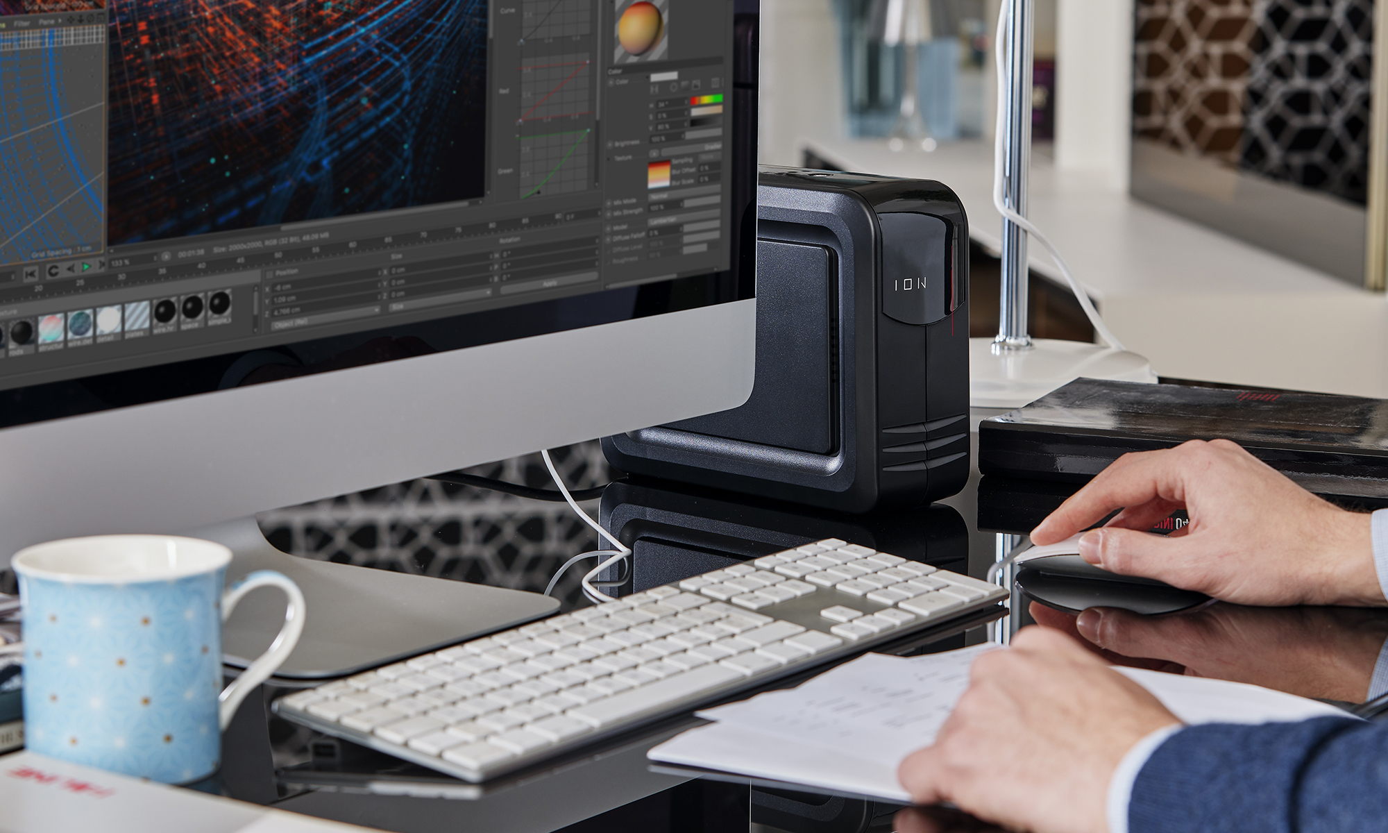 Una persona usa un computer a una scrivania. Un monitor mostra un'interfaccia software. C'è una tastiera, un mouse, una tazza e un dispositivo di archiviazione nero.