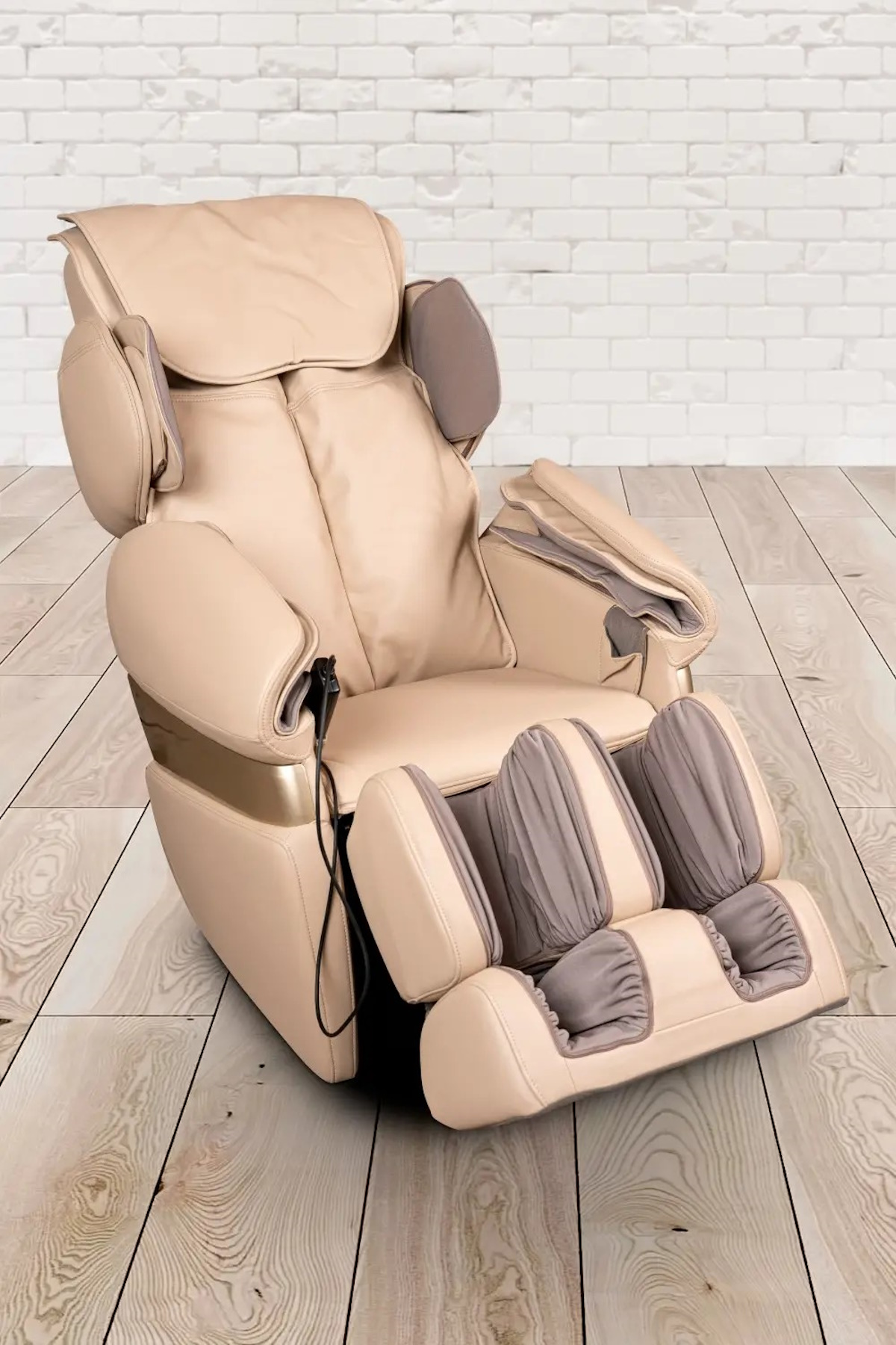 Ein beiger Massagesessel steht auf einem Holzboden. Der Hintergrund ist eine weiße Ziegelmauer. Der Stuhl hat eine Kopfstütze, Armlehnen und eine Fußstütze.