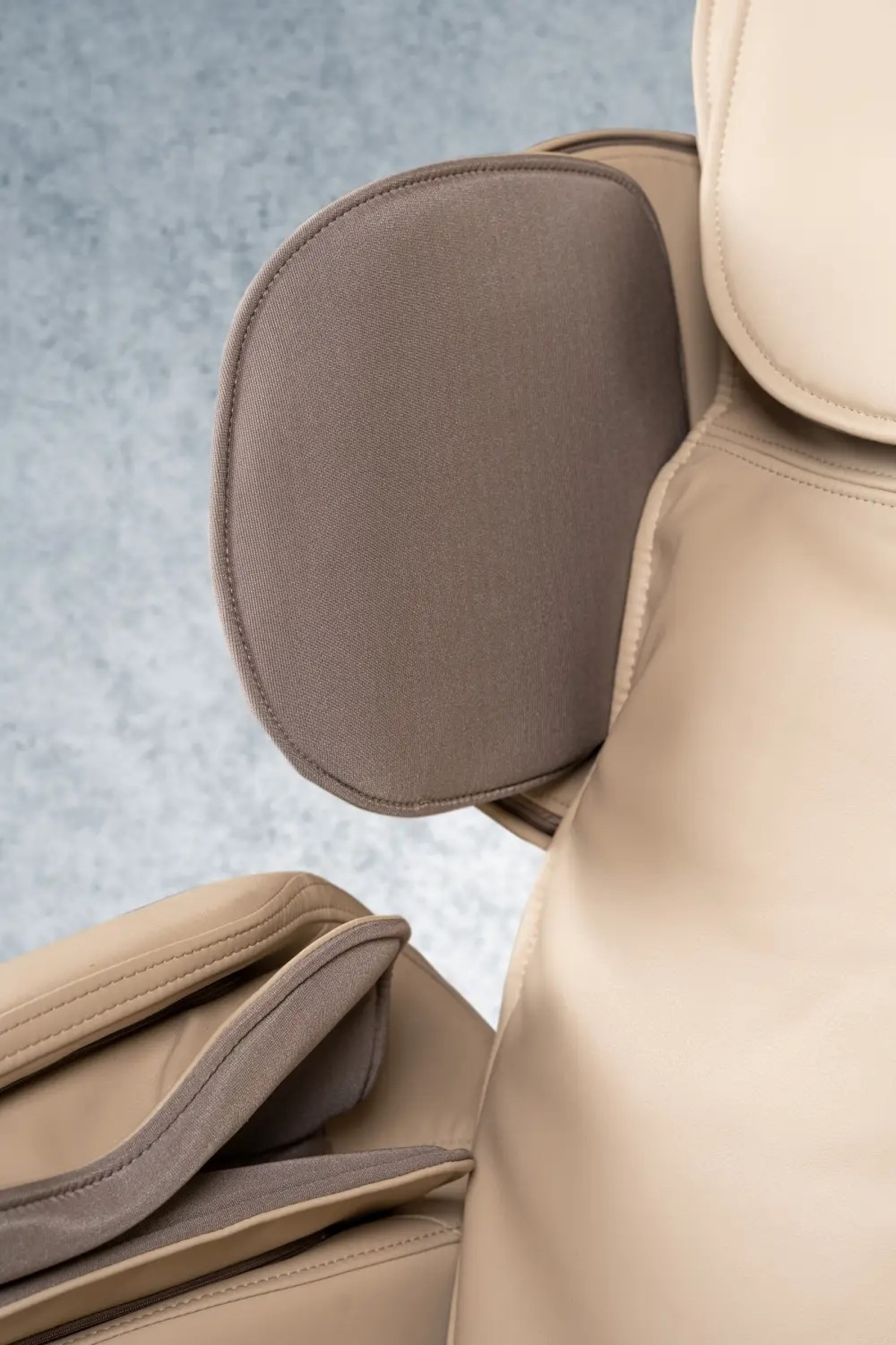 Nahaufnahme eines beigen Massagesessels mit grauer Kopfstütze. Der Stuhl besteht aus Leder und Stoff, vor grauem Hintergrund.