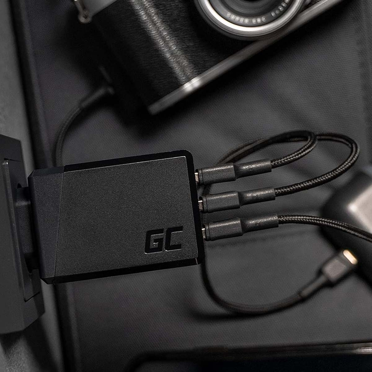 Cargador negro con tres cables enchufados. Una cámara y un estuche de carga en el fondo. El cargador está enchufado a la pared.