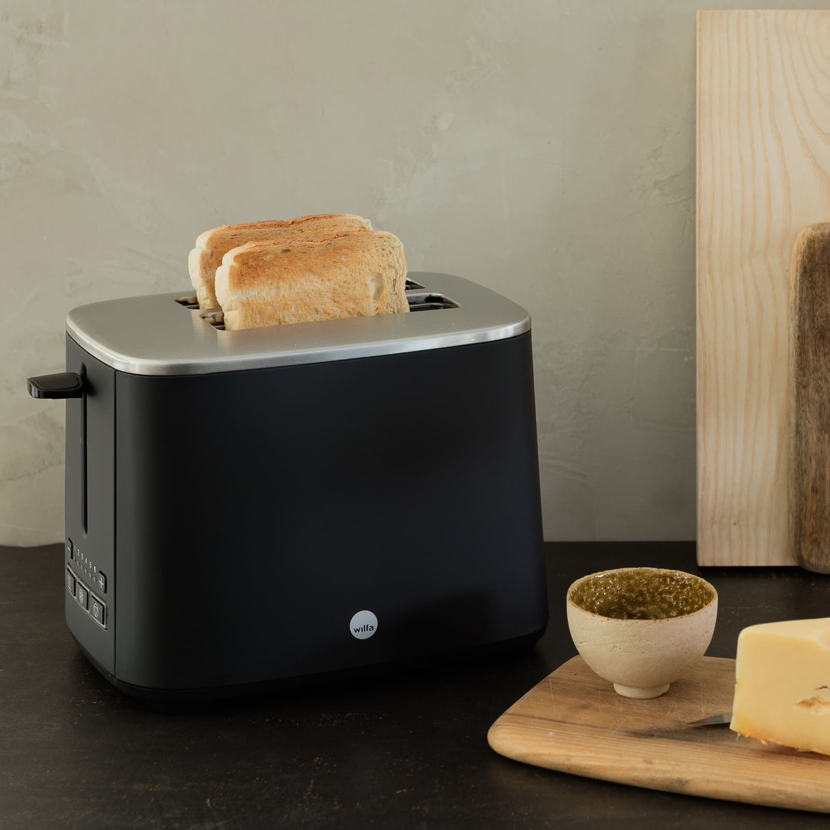Ein schwarzer Toaster mit zwei Toastscheiben. Eine Schüssel mit grünem Dip, Käse und Schneidebrettern stehen auf dem Tisch.
