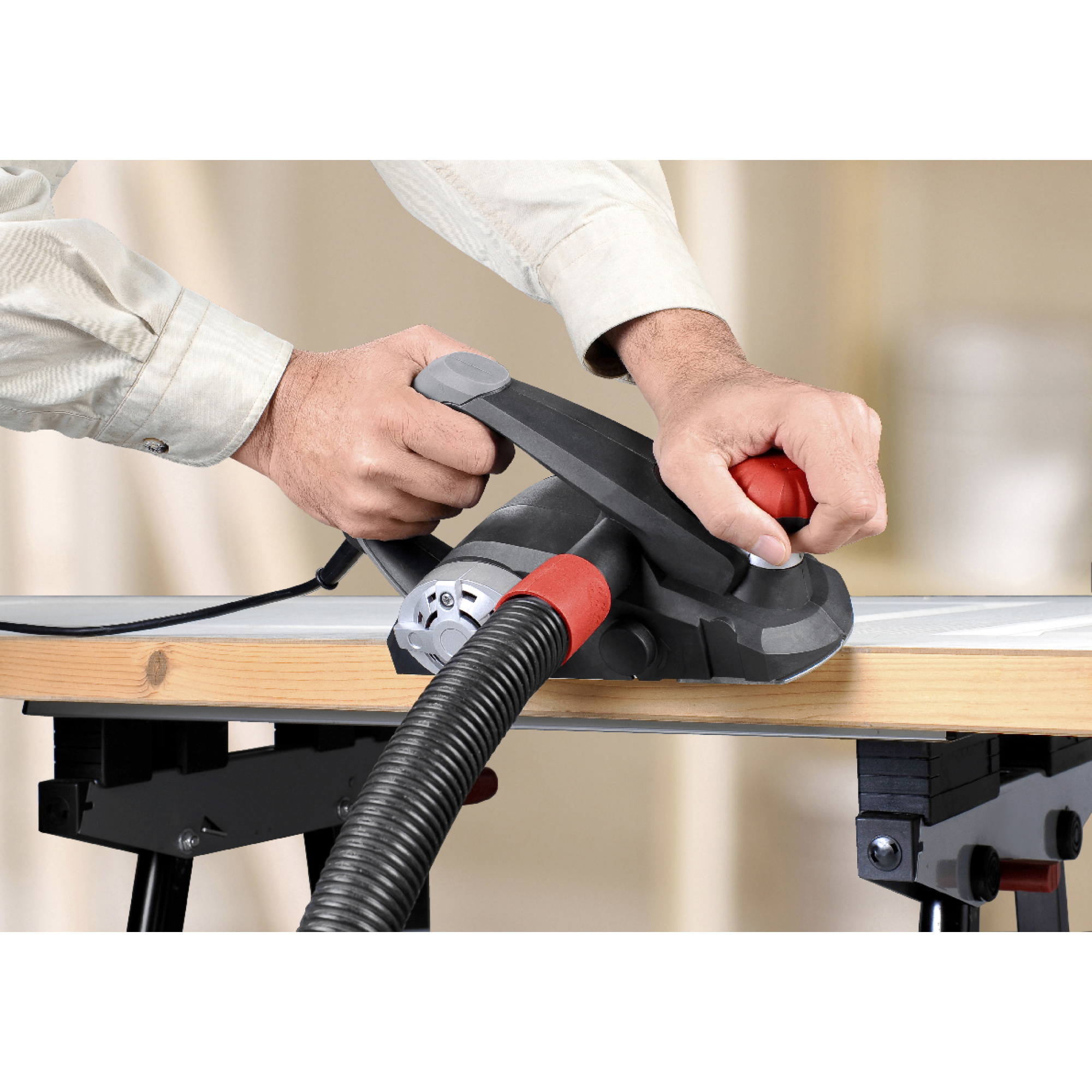 Osoba używa strugarki elektrycznej na drewnianej desce. Narzędzie jest czarne i szare z czerwonym uchwytem. Podłączony jest czarny wąż.