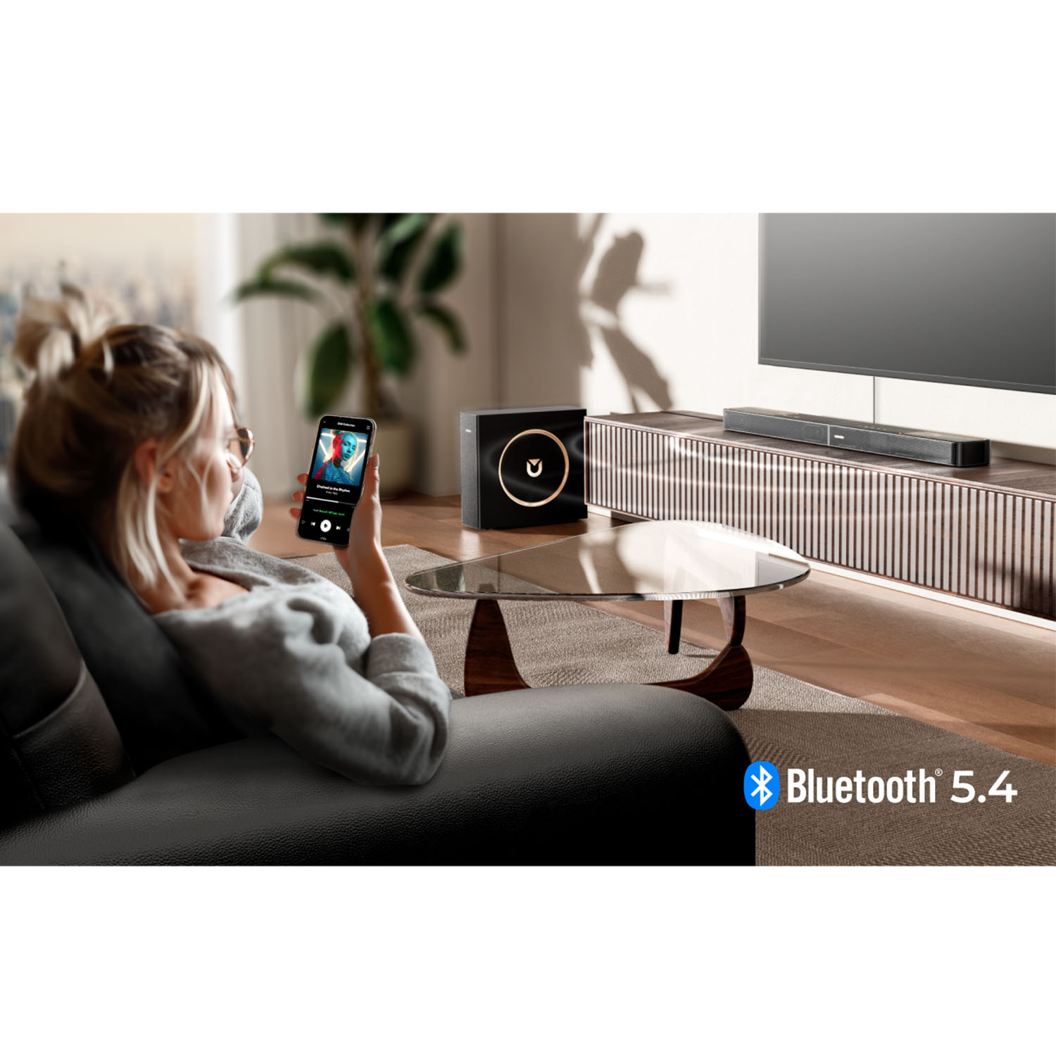 Kobieta na sofie używa telefonu do odtwarzania muzyki, głośnik i telewizor w tle. Logo Bluetooth 5.4.