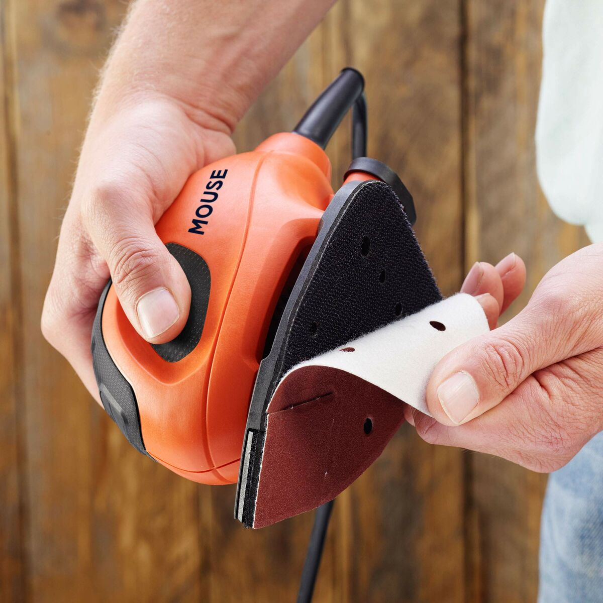 Une personne tient une ponceuse souris orange, changeant le papier de verre. Le fond est une surface en bois. La ponceuse a un câble noir.