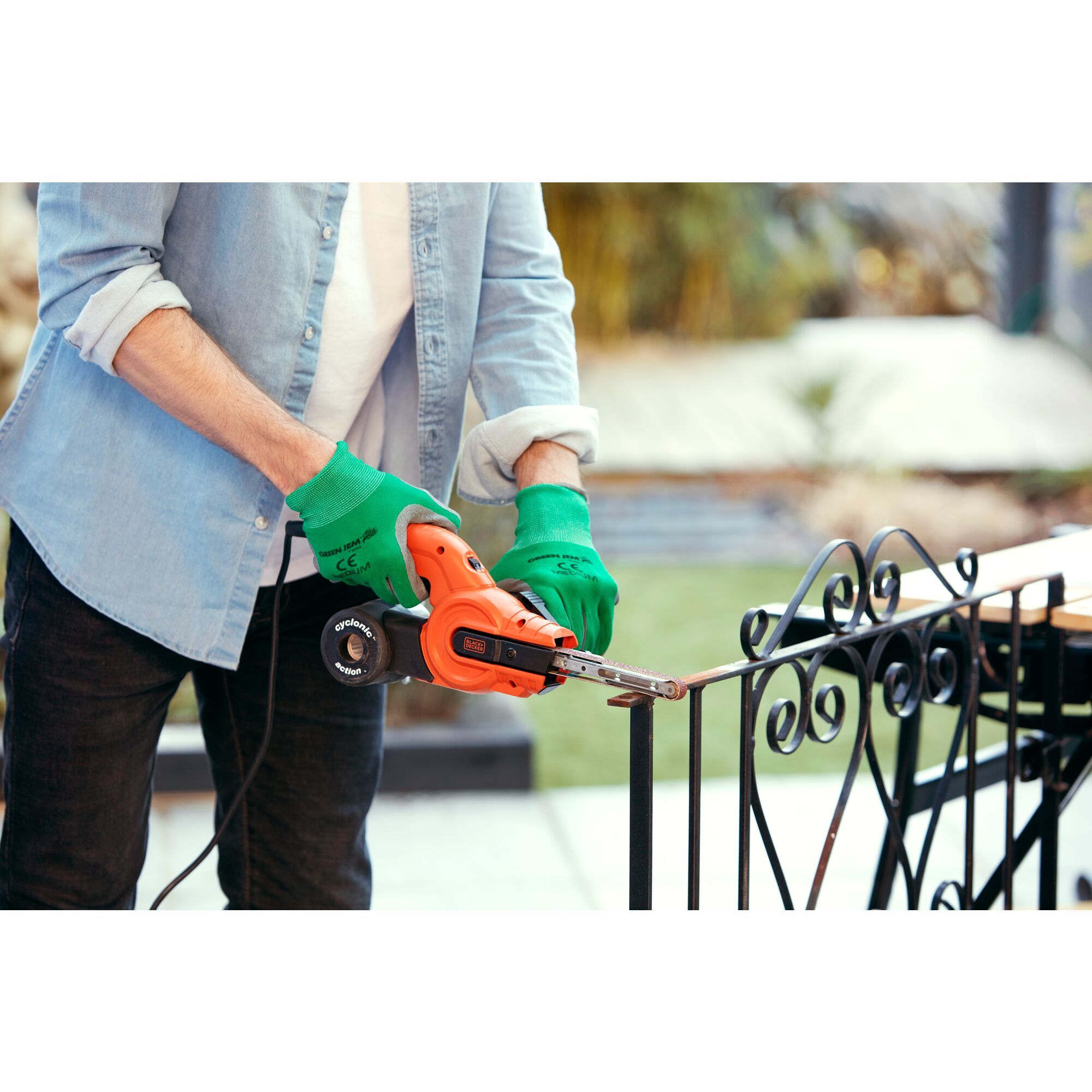 Person verwendet einen orangefarbenen Schleifer an einem Metallzaun. Grüne Handschuhe, blaues Hemd, schwarze Hose. Hintergrund ist verschwommenes Laub.