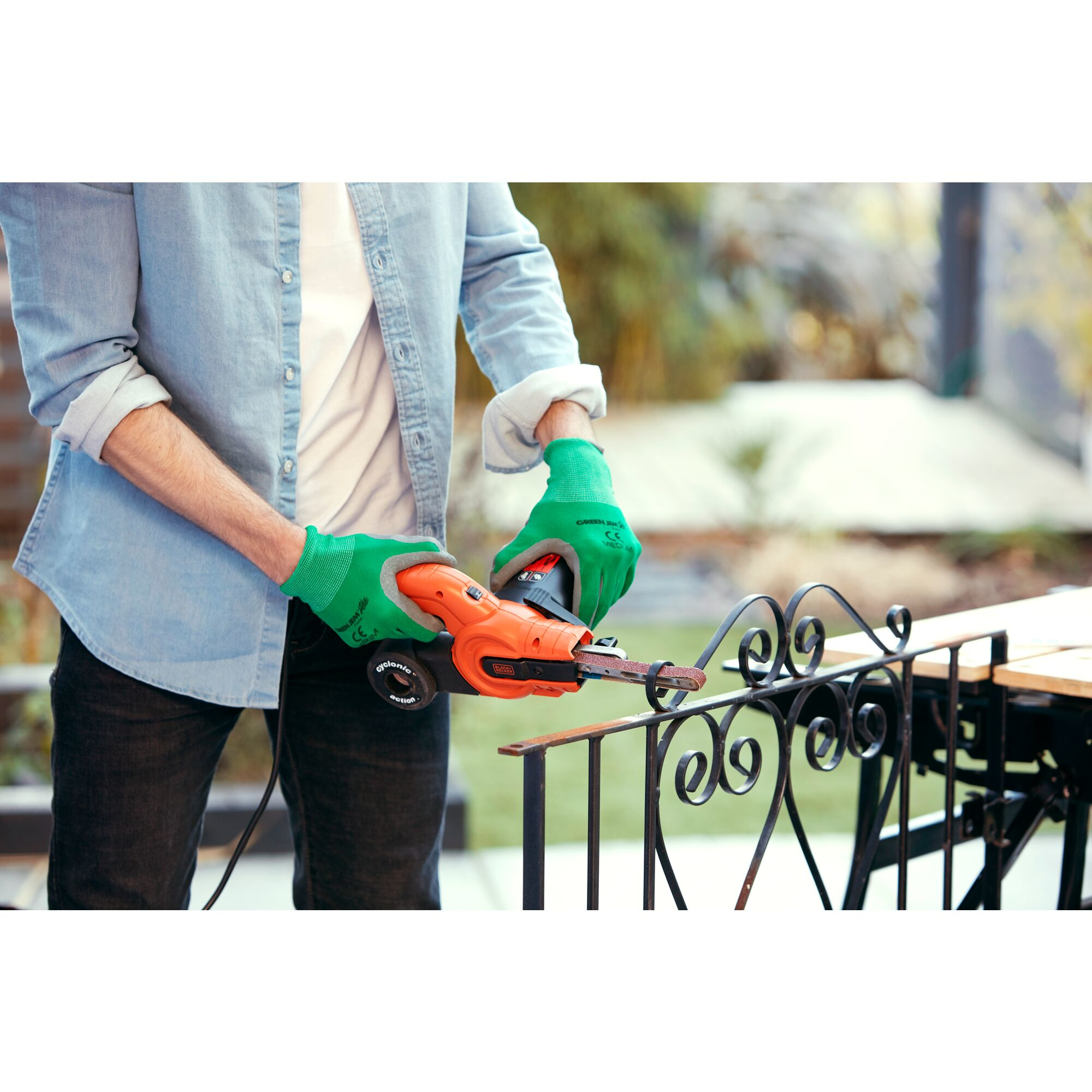 Eine Person mit grünen Handschuhen benutzt einen Schleifer an einem schmiedeeisernen Zaun. Die Person befindet sich in einem Garten.