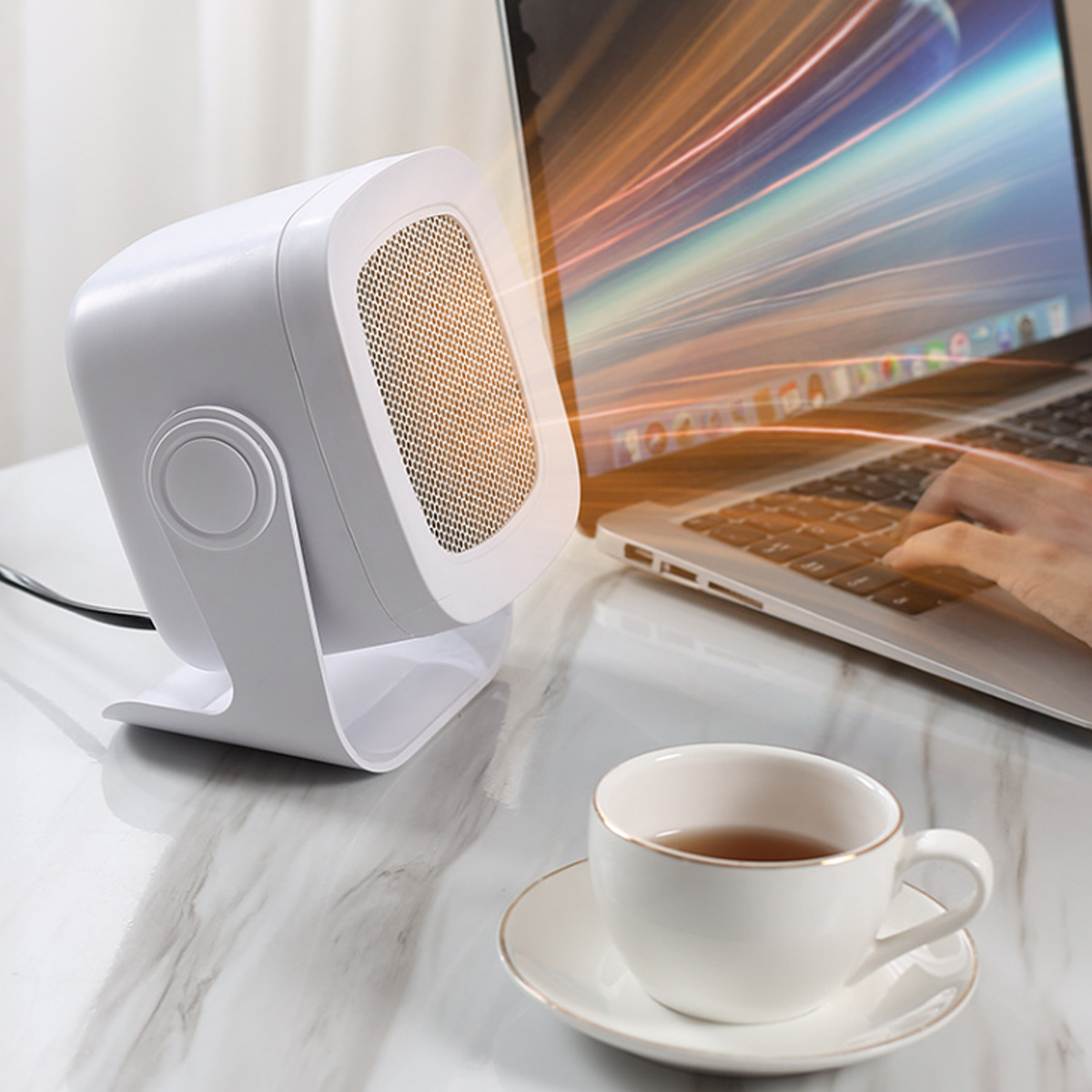Ein weißer Heizlüfter steht auf einem Schreibtisch neben einem Laptop und einer Tasse Tee. Eine Person tippt auf dem Laptop. Der Heizlüfter ist eingeschaltet.