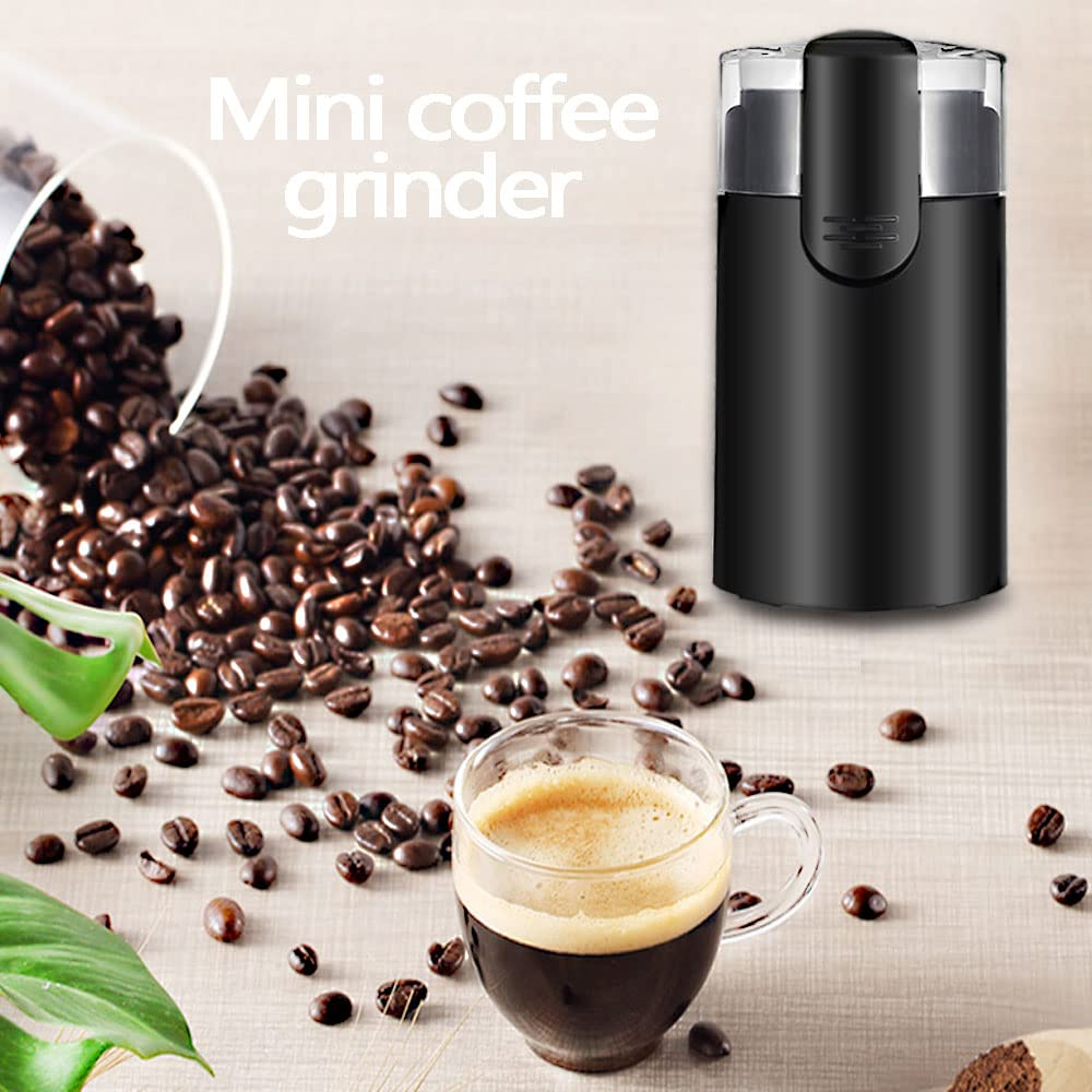 Eine schwarze Kaffeemühle, verstreute Kaffeebohnen, eine Tasse Kaffee mit Schaum und ein Blatt auf einer Holzoberfläche.