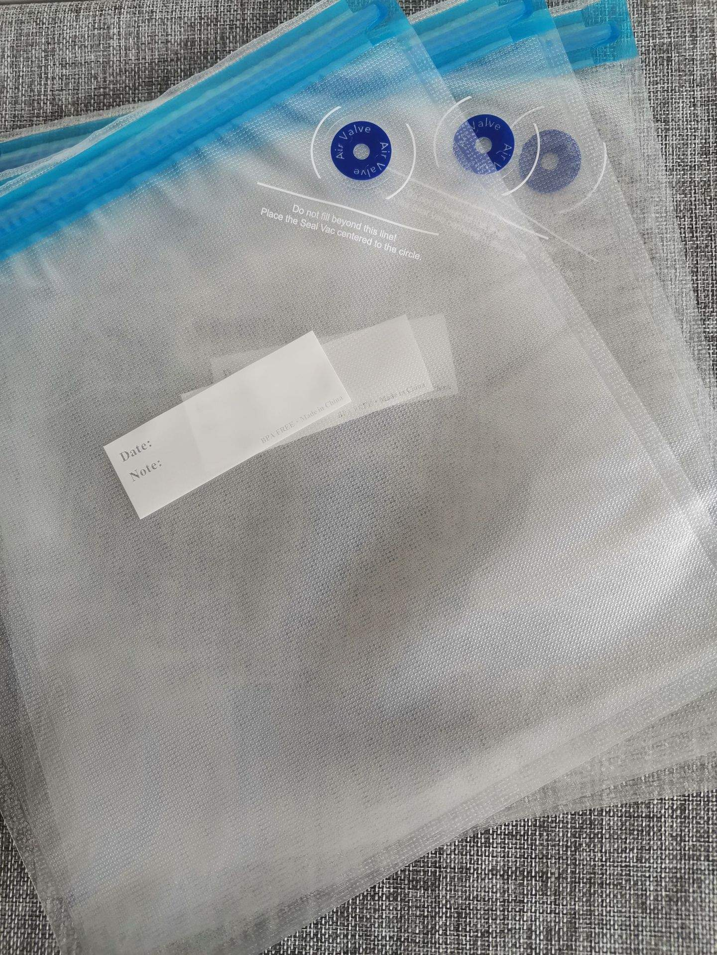 Transparente Vakuumbeutel mit blauen Akzenten. Text lautet 'Air Valve'. Hintergrund ist grauer Stoff.