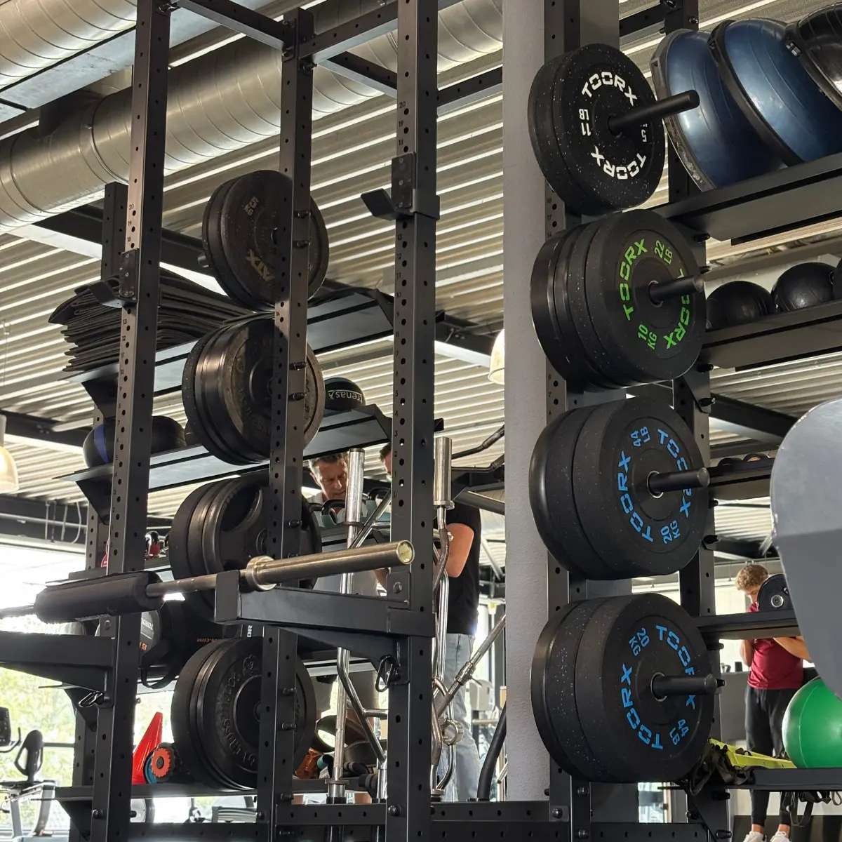 Gyminterieur met gewichtenrekken, diverse gewichten en mensen die aan het trainen zijn. Gewichten zijn zwart met gekleurde markeringen. Apparatuur zichtbaar.