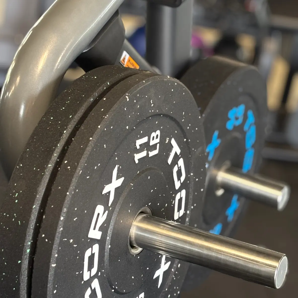 Close-up van halterschijven op een gymrek. Zwarte schijven met witte letters, zilveren stang. De achtergrond toont meer fitnessapparatuur.