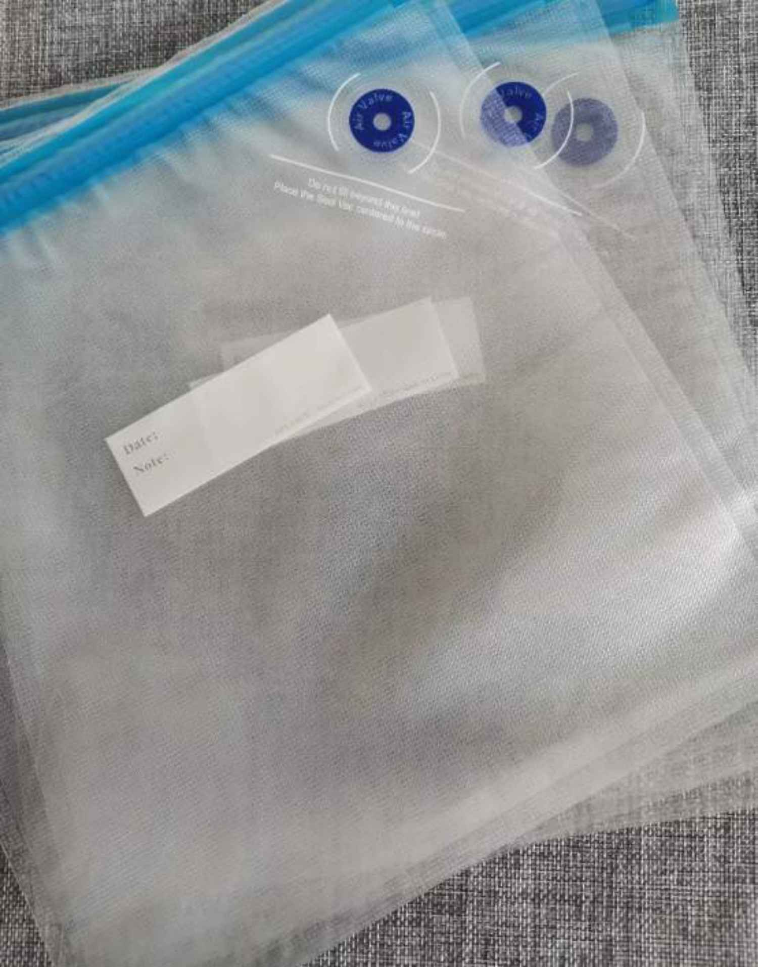 Transparente Vakuum-Aufbewahrungsbeutel mit blauen Akzenten, Etiketten und Luftventil. Die Beutel sind auf einer strukturierten, grauen Oberfläche gestapelt.