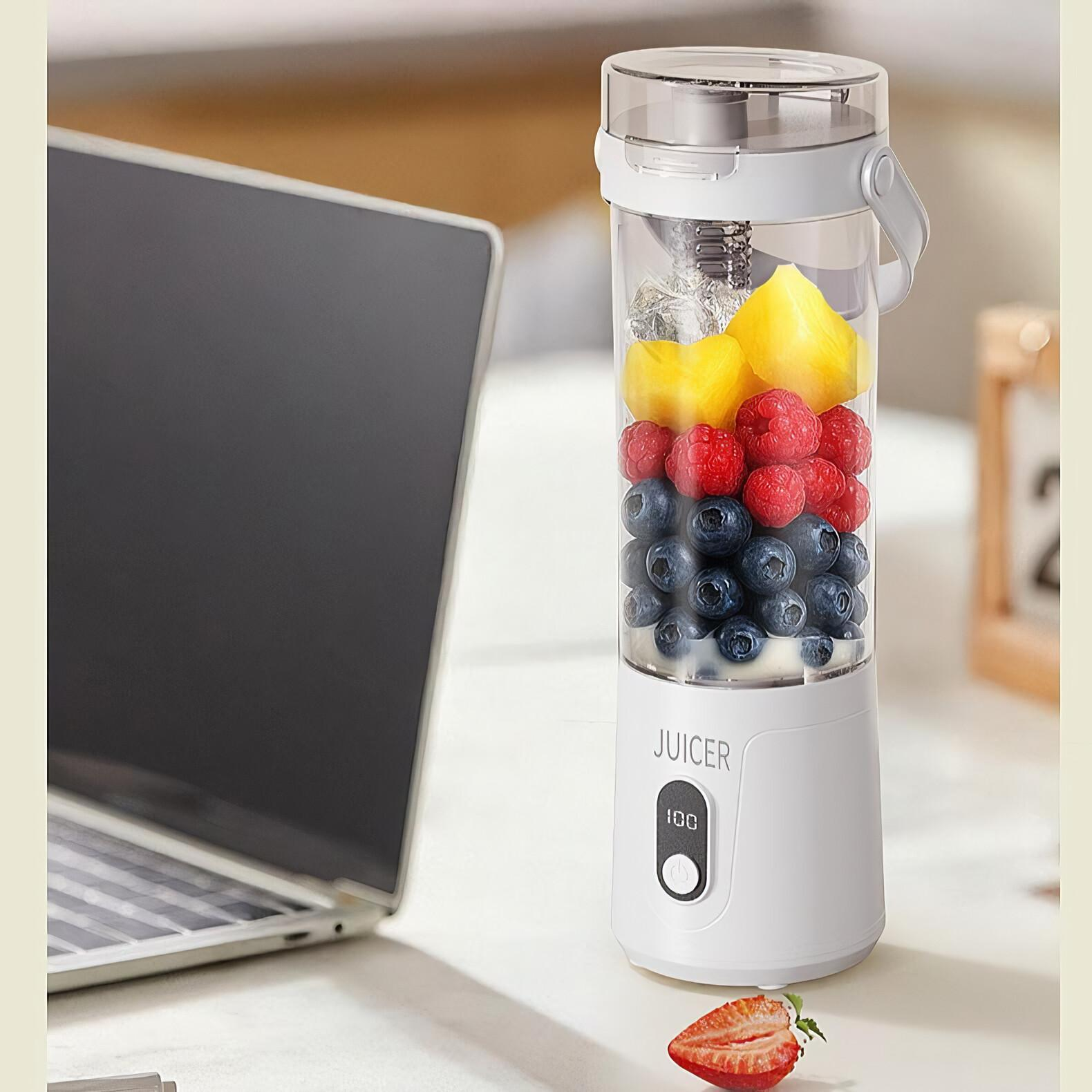 Ein weißer Entsafter mit Früchten steht neben einem Laptop. Darunter liegt eine geschnittene Erdbeere. Früchte sind Blaubeeren, Himbeeren und Ananas.