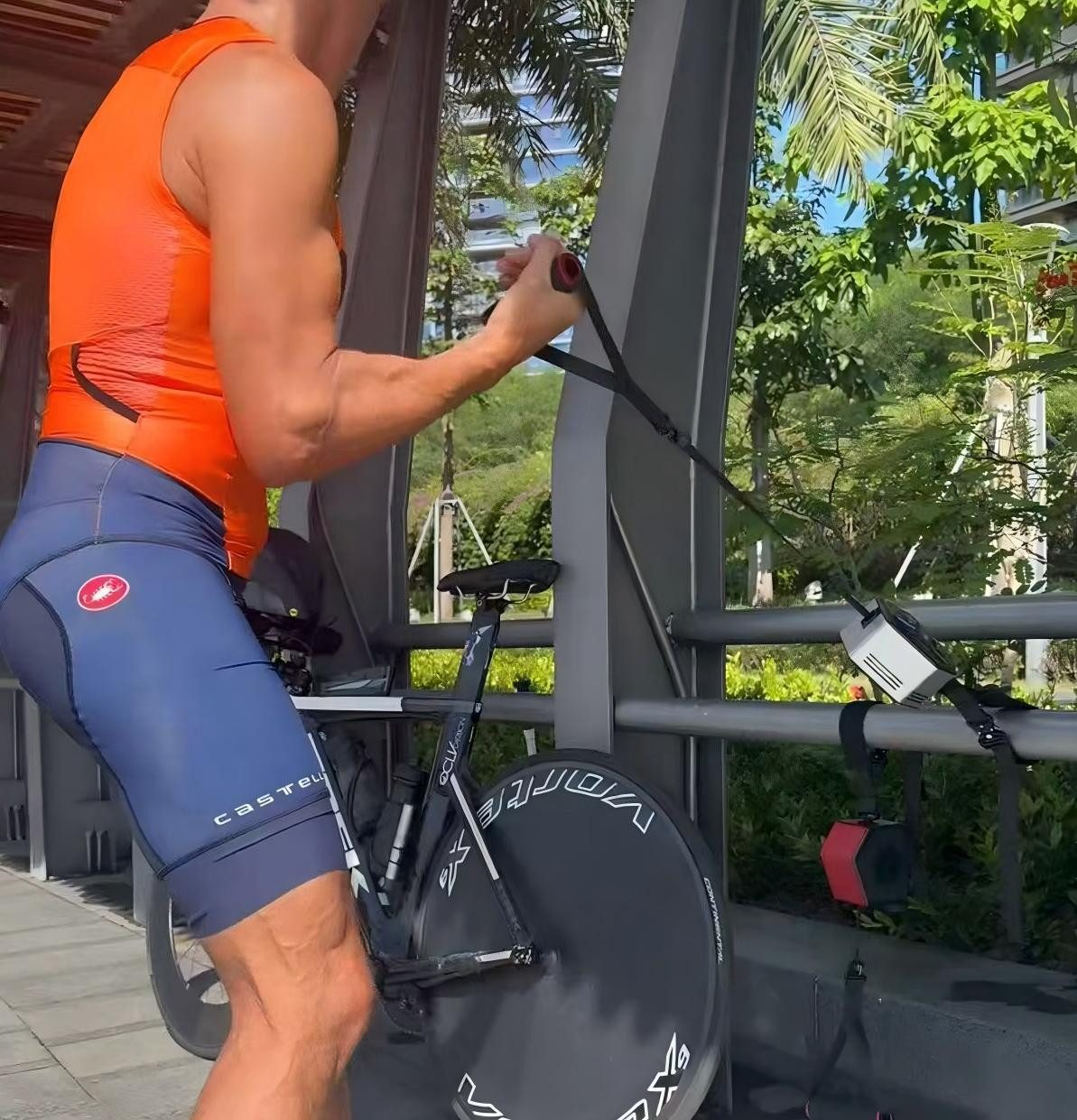 Ein Mann in orangefarbenem Oberteil und blauen Shorts trainiert im Freien mit einem Fahrrad und zieht ein Widerstandsband. Grüne Bäume sind im Hintergrund.