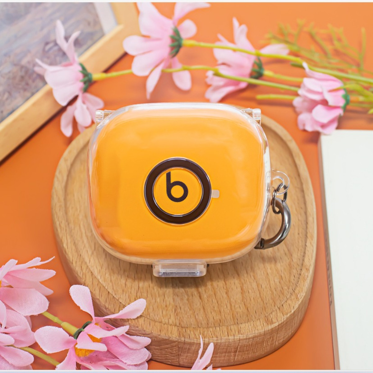 Orangenes Beats-Kopfhörer-Etui auf einem Holztablett, umgeben von rosa Blumen und Grün. Das Etui hat ein schwarzes "b"-Logo.