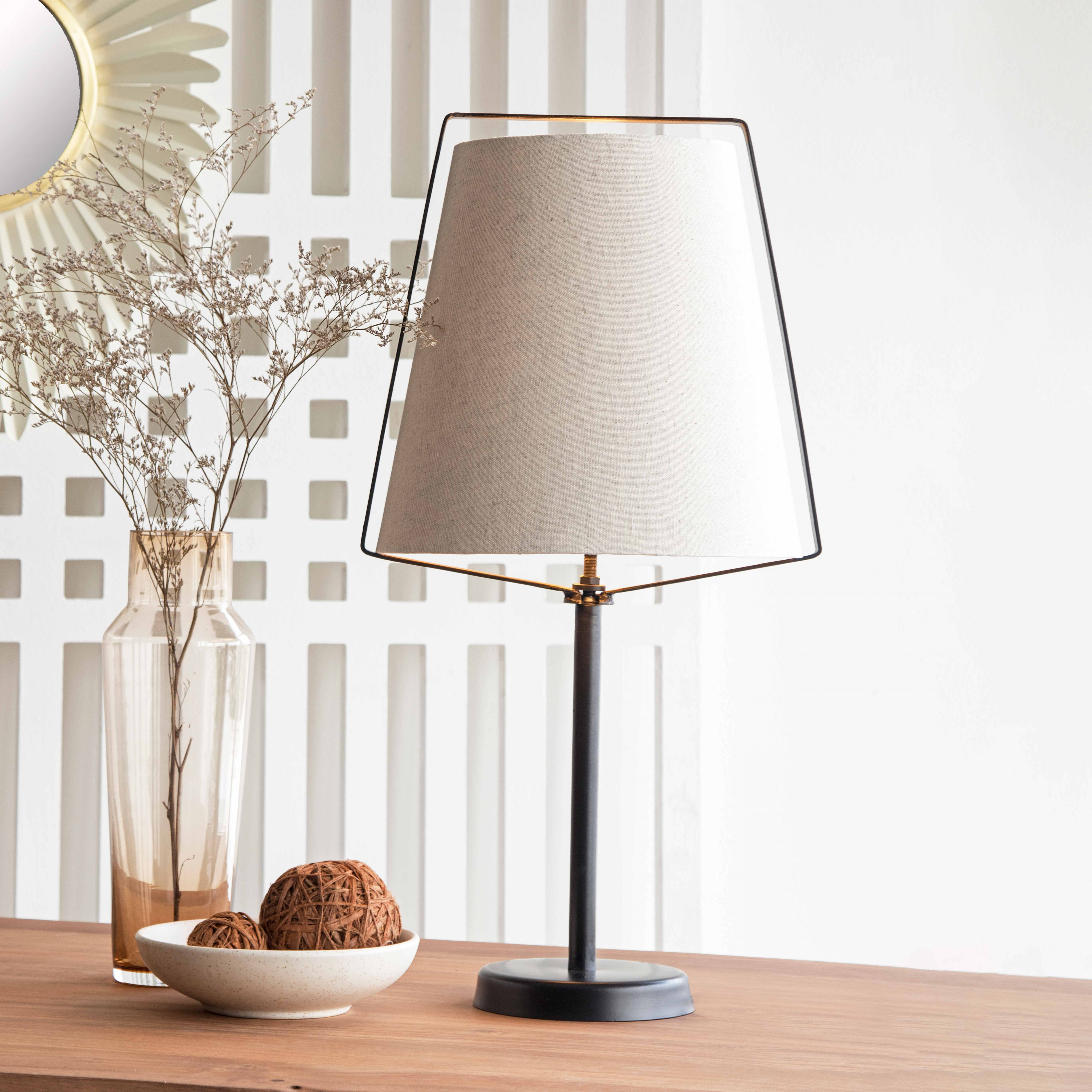 Eine Lampe mit beigem Schirm steht auf einer Holzoberfläche. Eine Vase und Zierkugeln sind ebenfalls vorhanden.