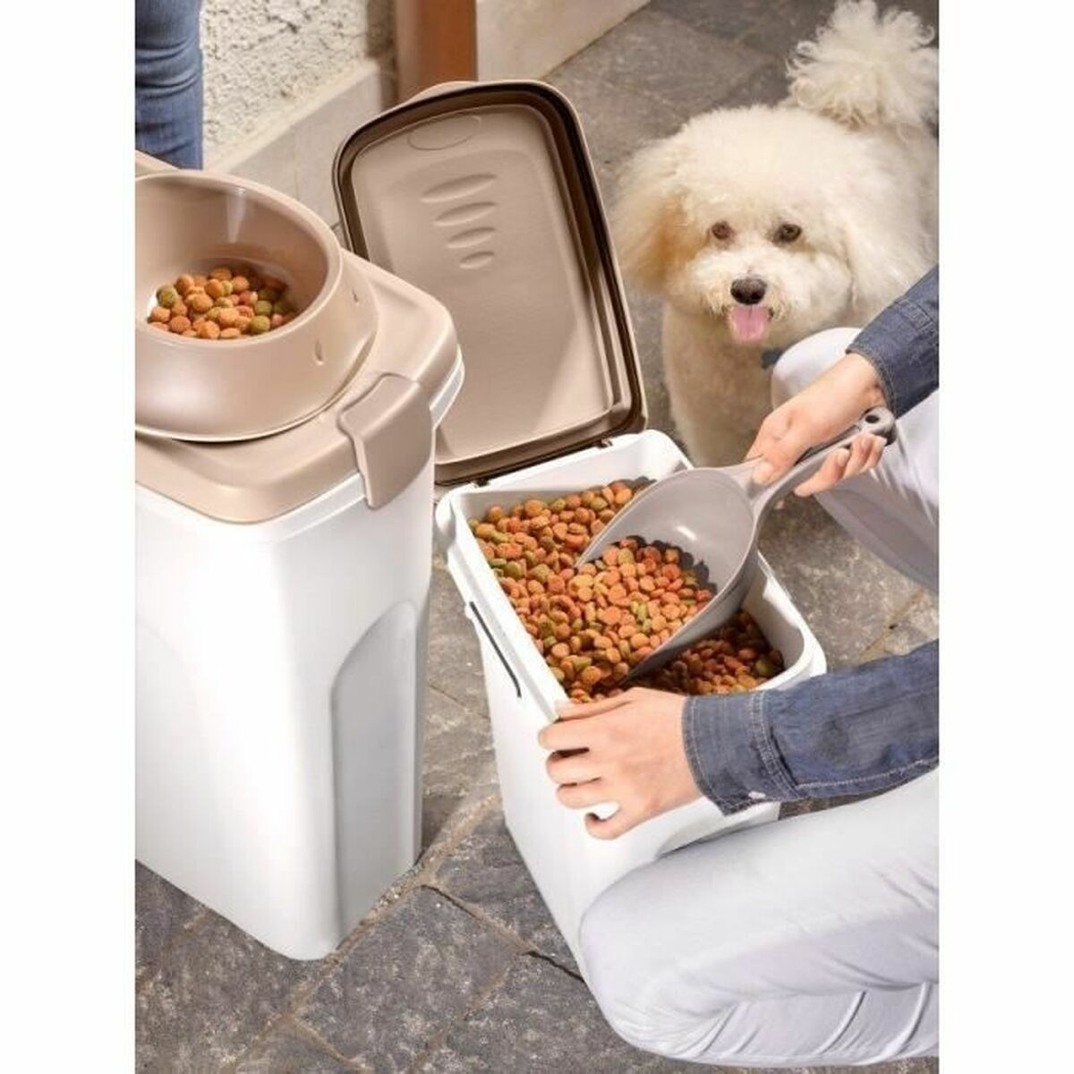 Una persona versa cibo per cani in una ciotola, il cane osserva. Contenitori pieni di cibo secco per cani sono vicini. Il cane ha il pelo bianco.