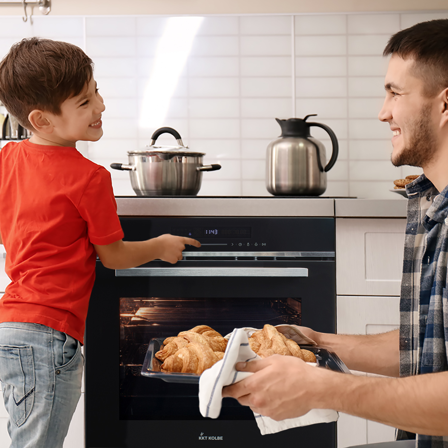 Ein Junge und ein Mann sind in einer Küche. Der Mann hält ein Blech mit Croissants, während der Junge auf den Ofen zeigt.