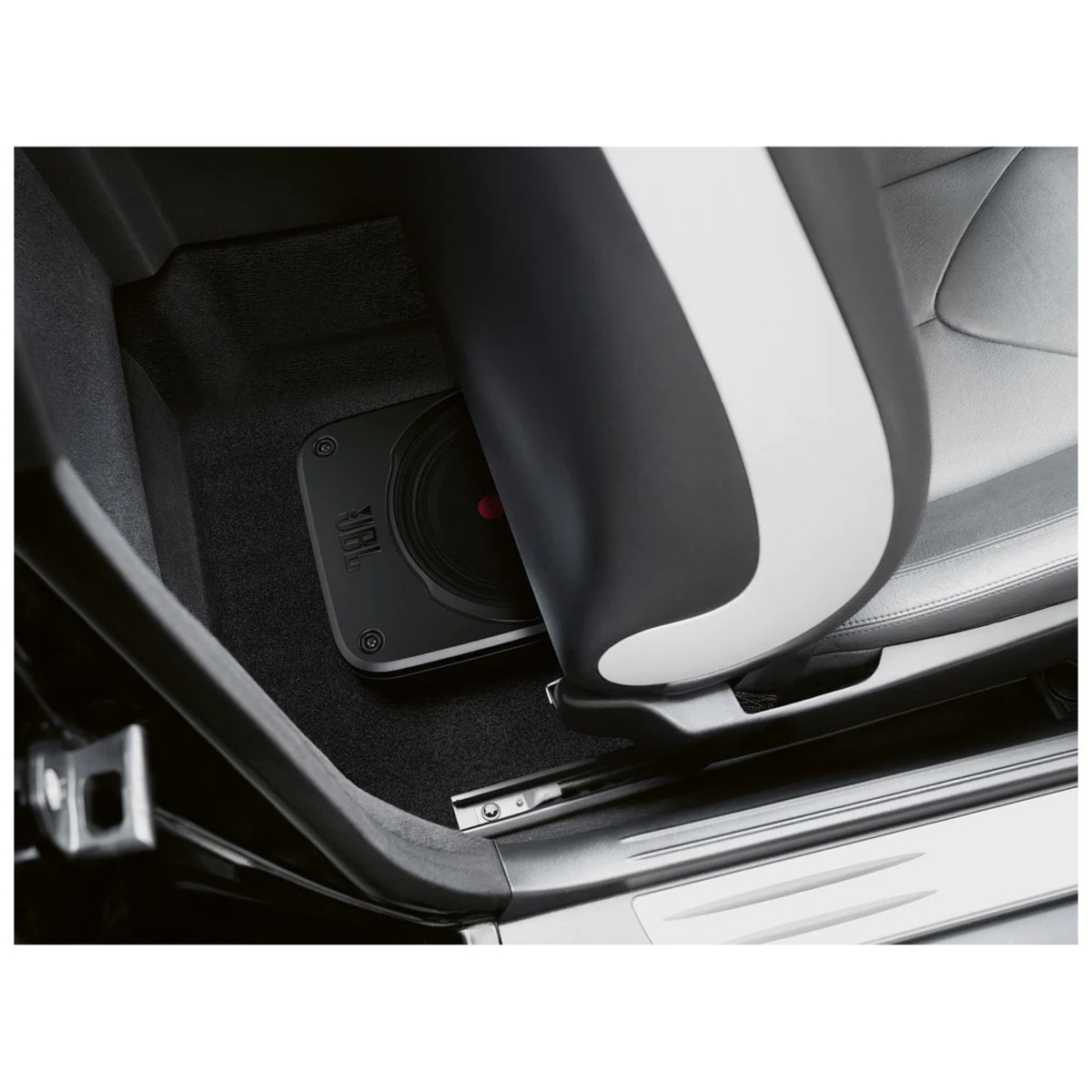 Autoinnenraum mit einem JBL-Subwoofer unter dem Sitz. Schwarz-weißer Sitz, schwarzer Teppich und Türrahmen sichtbar.
