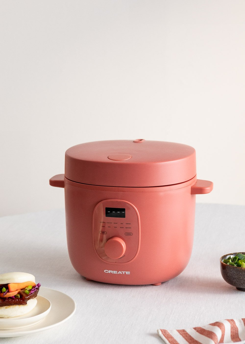 Una olla de cocción múltiple de color coral está centrada. Un pequeño bao con verduras está en un plato. Un cuenco con cebolletas está a la derecha.