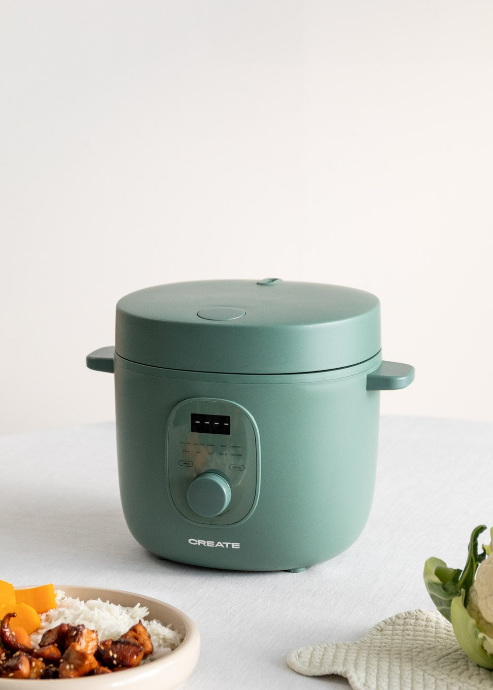 Una olla a presión eléctrica turquesa sobre un mantel blanco. Comida y coliflor a los lados. El fondo es blanco liso.