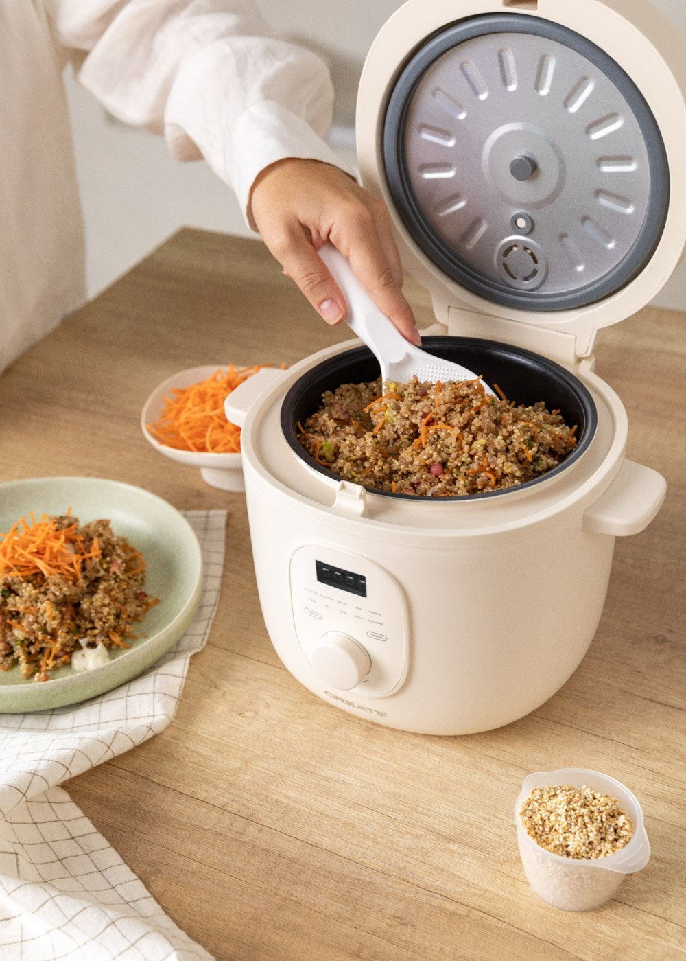 Una persona saca comida de una arrocera. Quinoa cocida con zanahorias está en la arrocera y en un plato. Un pequeño recipiente contiene granos.