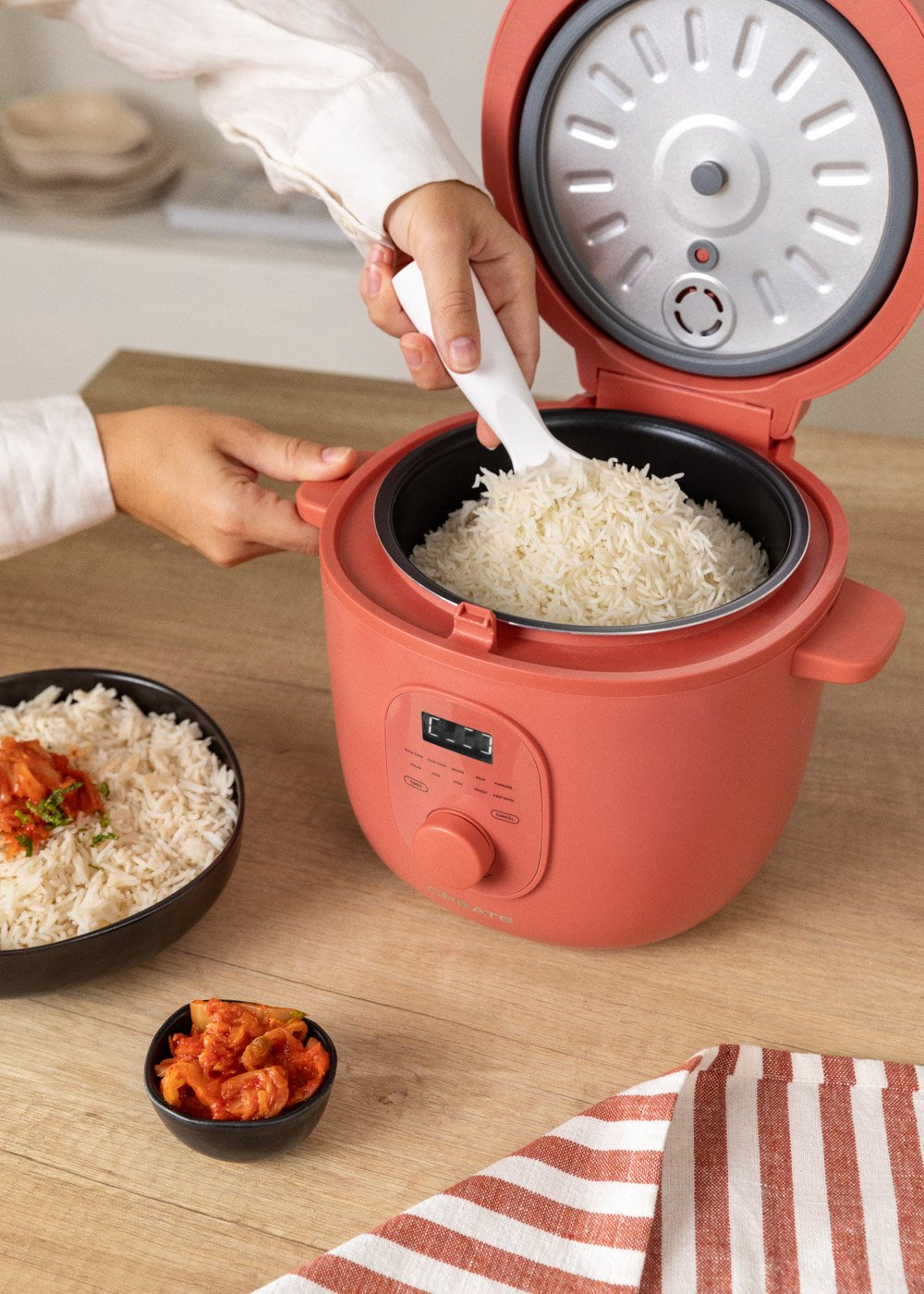 Una persona sirve arroz de una arrocera coral. Cuencos de arroz y kimchi están cerca sobre una superficie de madera. Hay un paño a rayas.