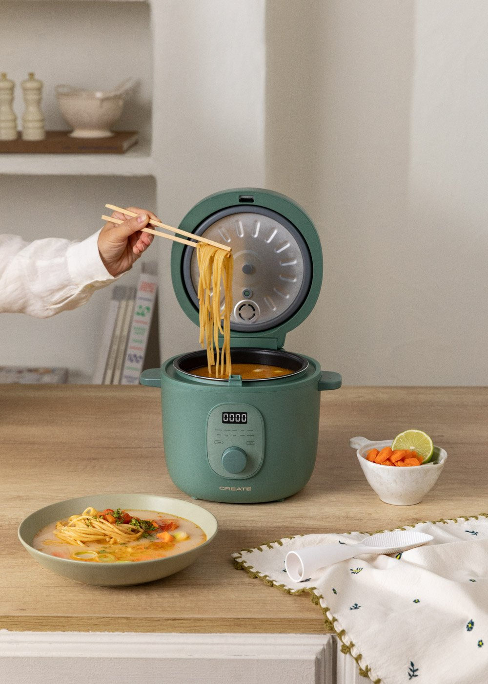 Una persona usa palillos para levantar fideos de una olla verde. Un cuenco de sopa y zanahorias con lima están en la escena.