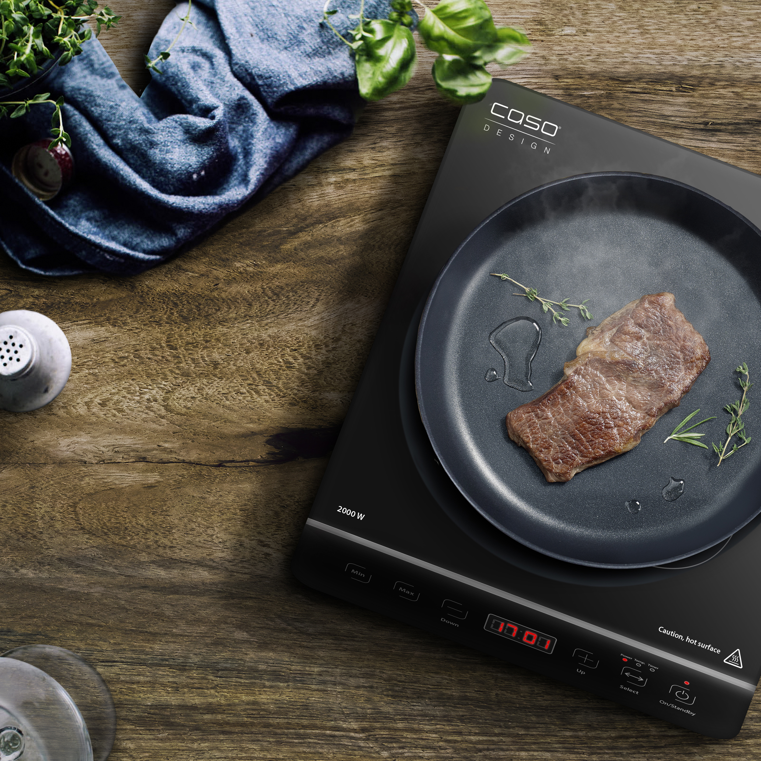 Una bistecca sta cuocendo in una padella su un piano cottura a induzione. Sono presenti anche erbe, un panno blu e una superficie in legno.