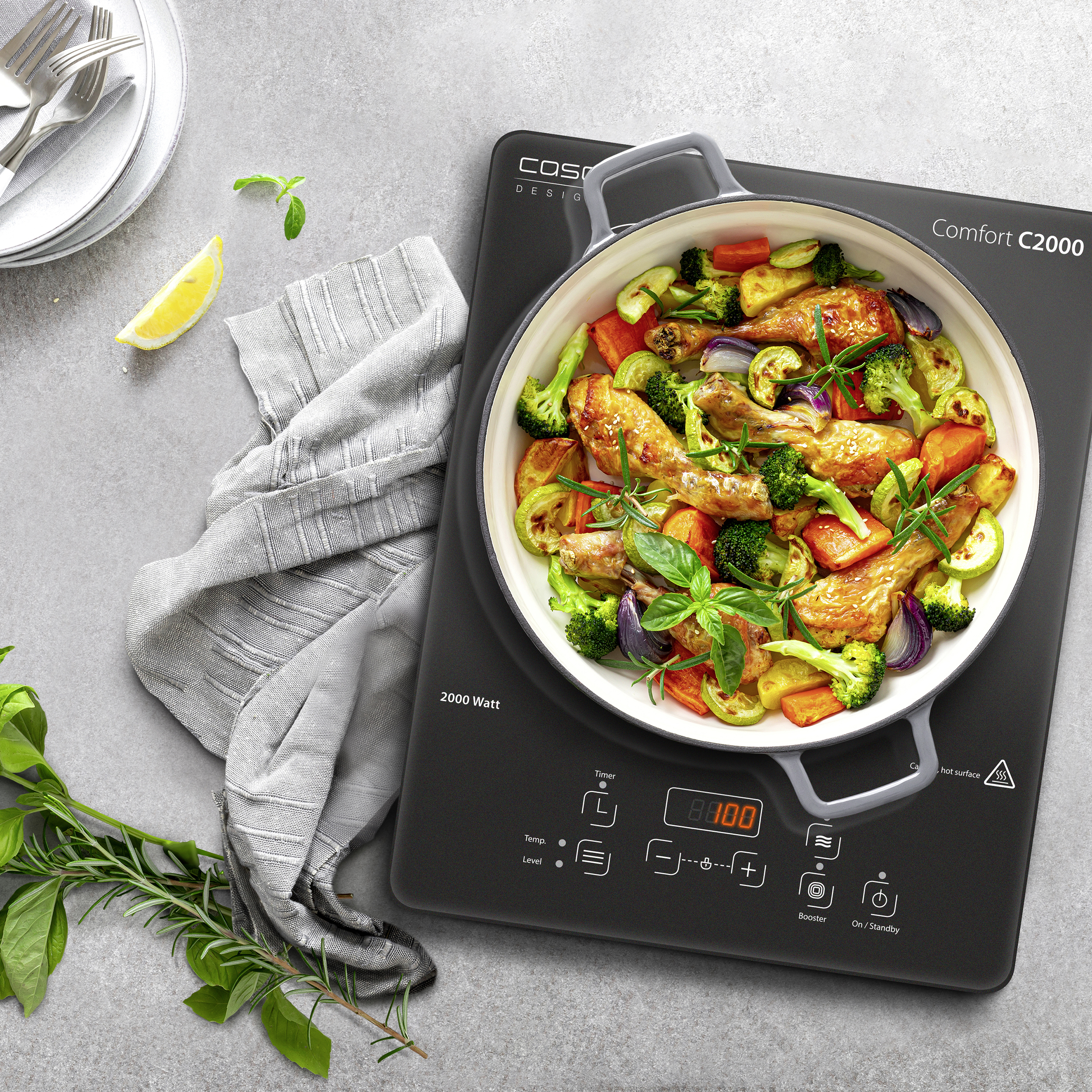 Une poêle avec du poulet et des légumes cuits sur une table de cuisson à induction noire. Assiettes et citron à gauche.