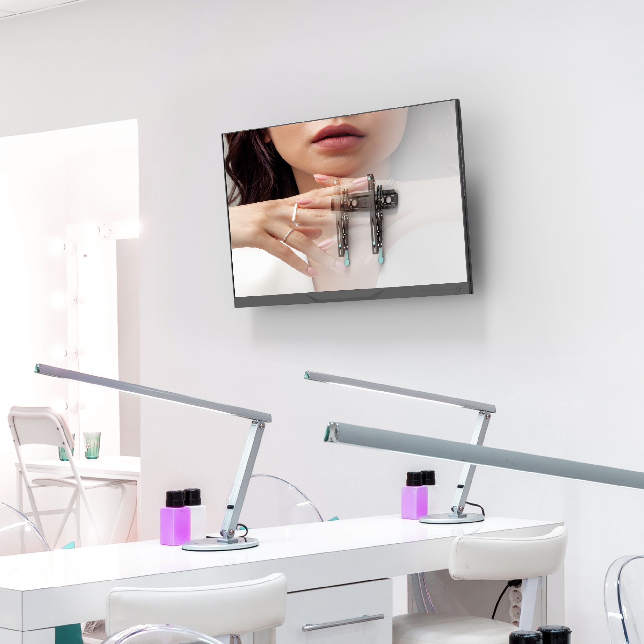 Wnętrze salonu paznokci z białymi stołami, krzesłami i lampami. Telewizor pokazuje dłoń z manicure. Różowe butelki i lustro w tle.