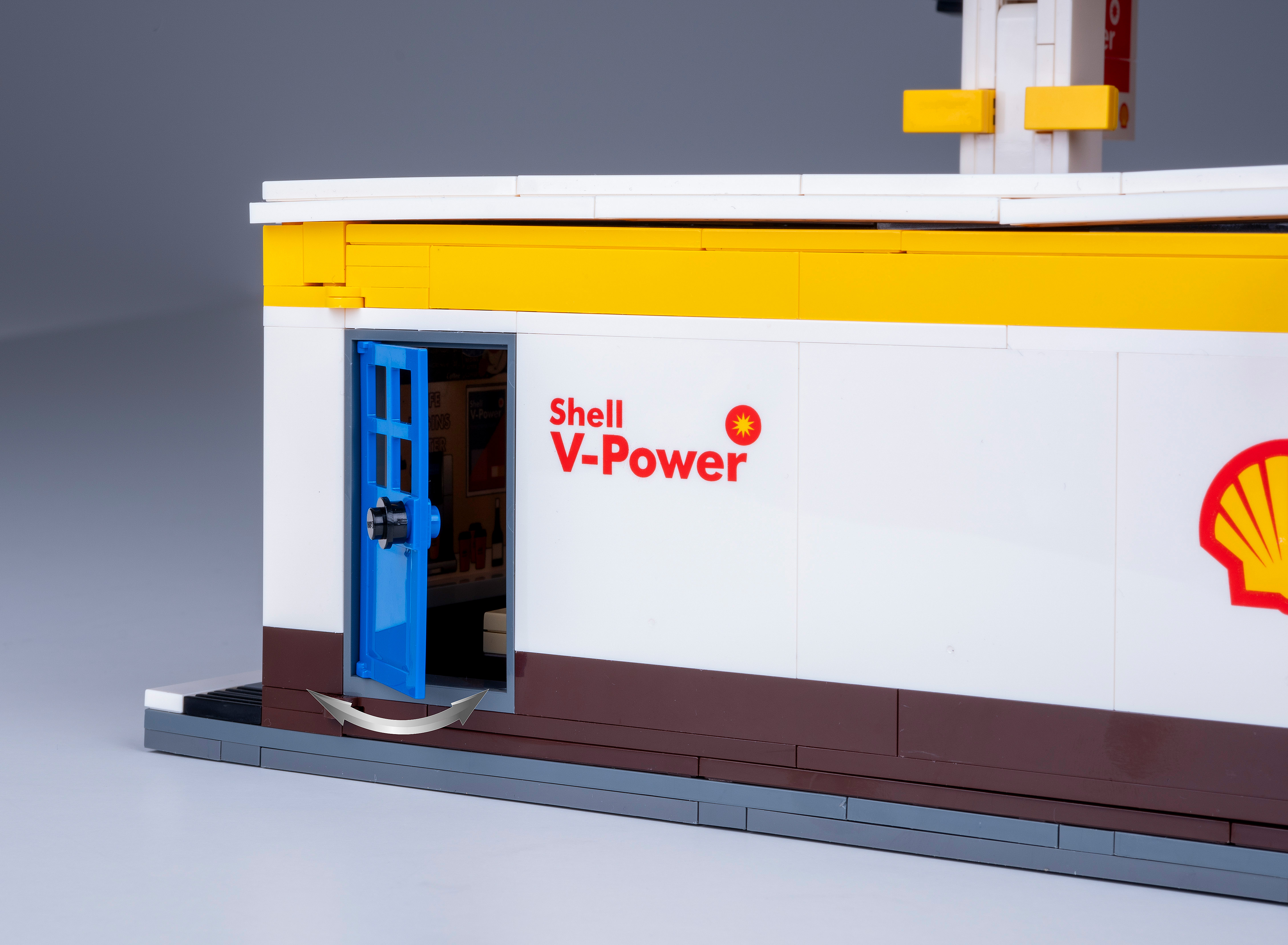 Lego Shell Tankstelle mit offener blauer Tür. Shell V-Power Logo sichtbar. Gelbes und weißes Gebäude mit grauem Sockel.