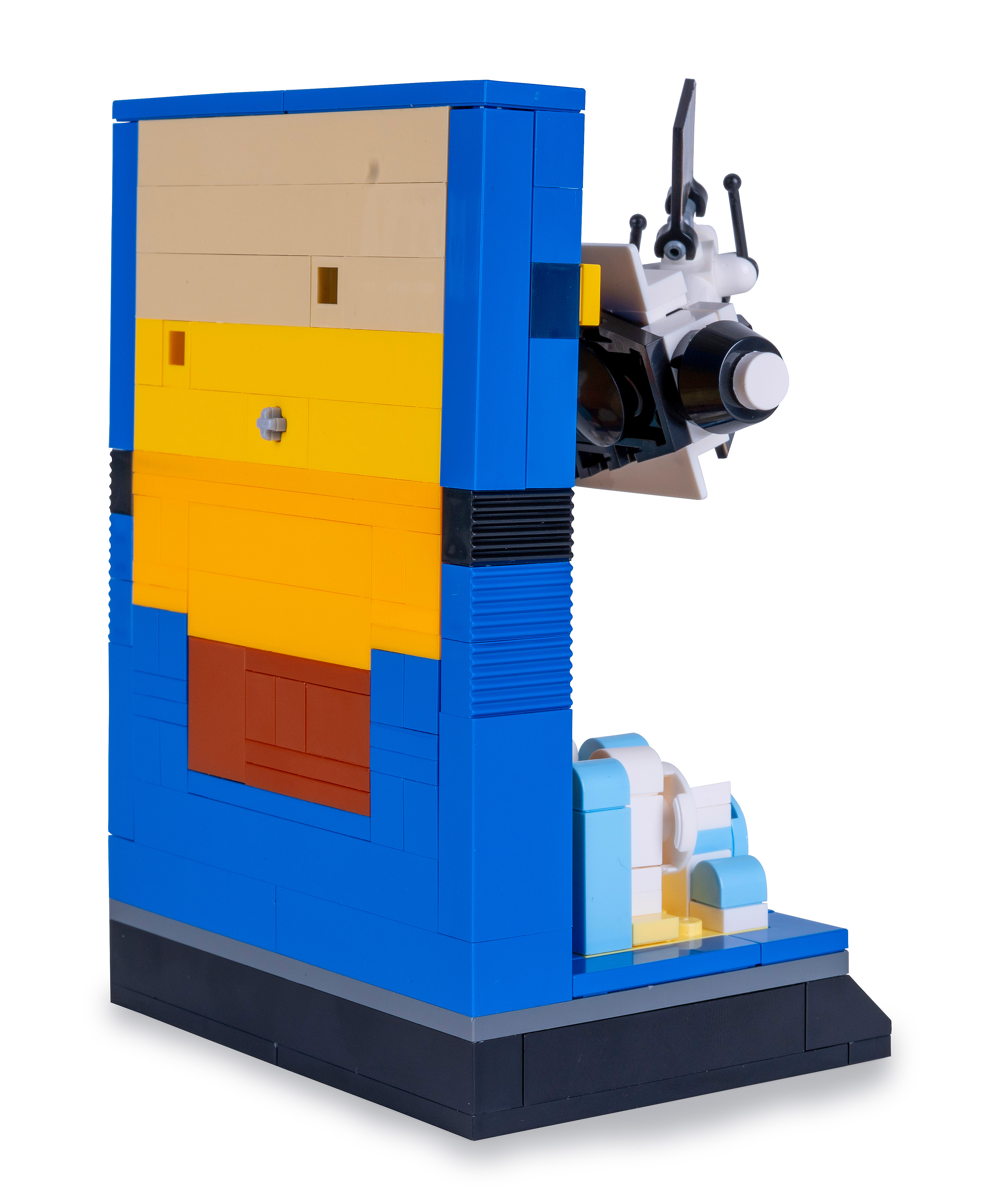 Ein LEGO-Modell einer Wand mit einer Portal-Kanone. Die Wand hat blaue, beige, gelbe und braune Blöcke. Die Kanone ist weiß und schwarz.