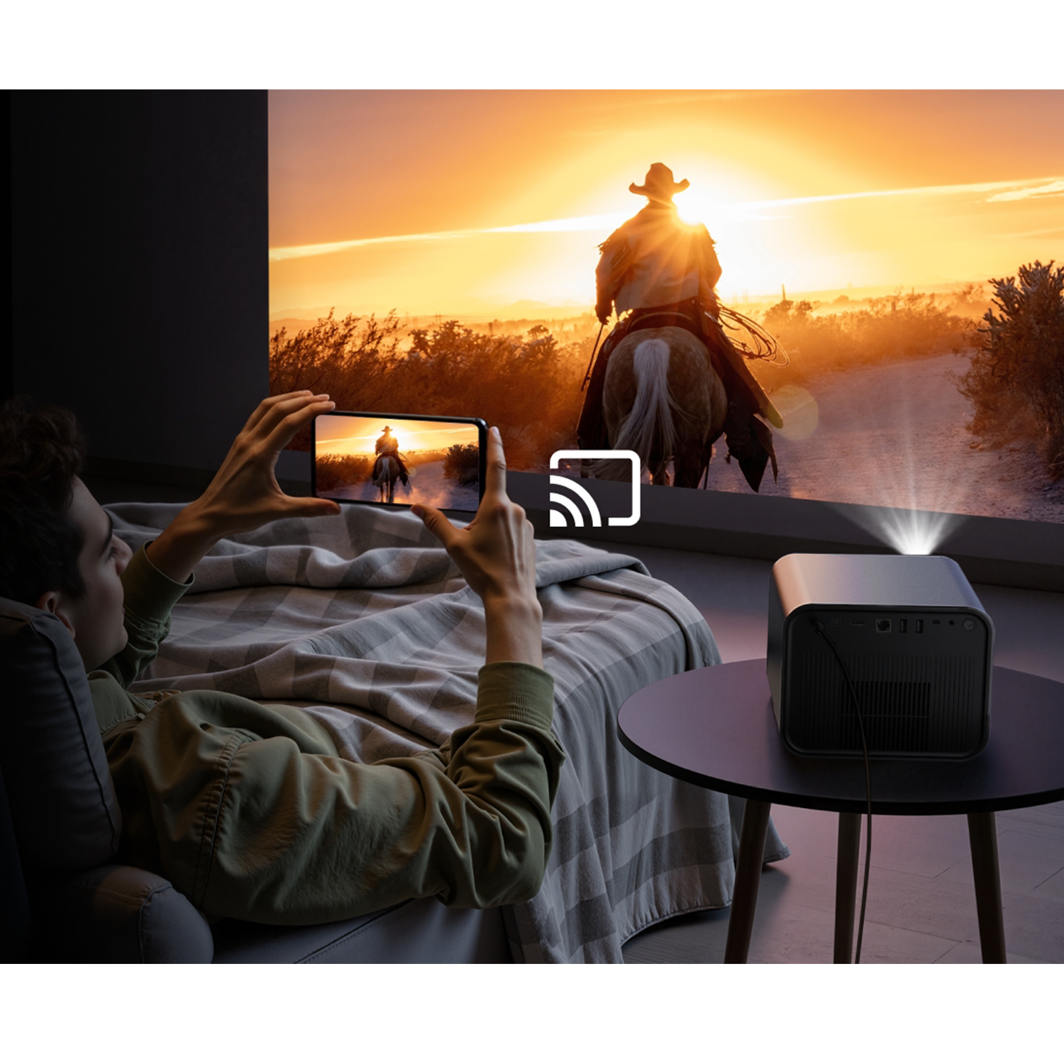 Eine Person liegt auf einem Bett und hält ein Telefon, das ein projiziertes Bild eines Cowboys auf einem Pferd bei Sonnenuntergang spiegelt. Ein Projektor steht auf dem Tisch.