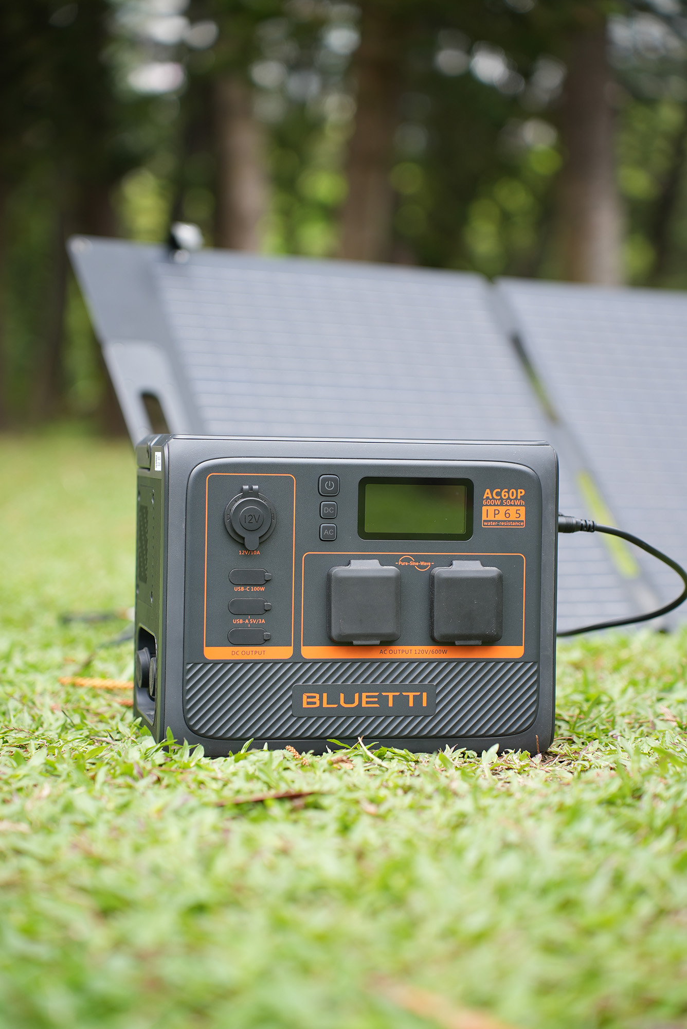Czarno-pomarańczowa stacja zasilania z napisem 'BLUETTI' stoi na trawie z panelem słonecznym w tle. Drzewa są rozmyte.