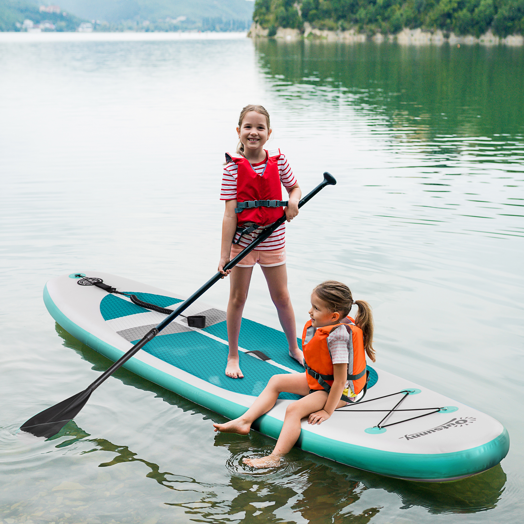 Dos niñas en una tabla de paddle en un lago. Una sostiene un remo, la otra está sentada. Ambas llevan chalecos salvavidas. Montañas al fondo.
