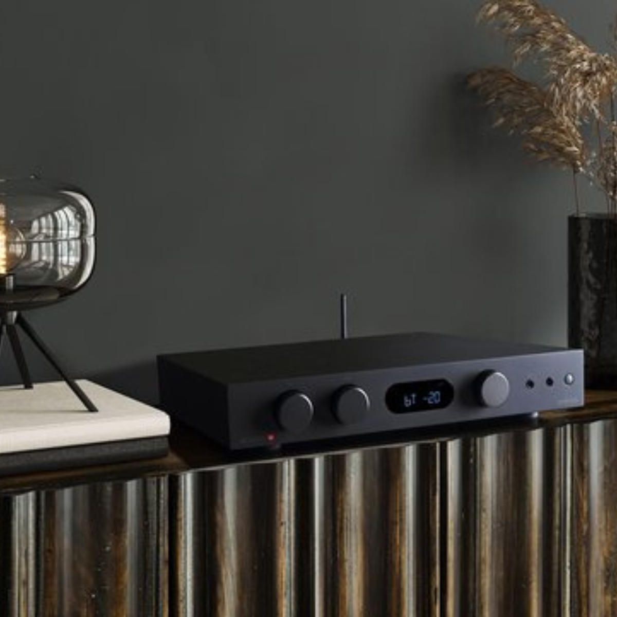 Een zwarte audioversterker op een houten kast met een lamp en gedroogde bloemen. De versterker heeft knoppen en een display met 'bt -20'.