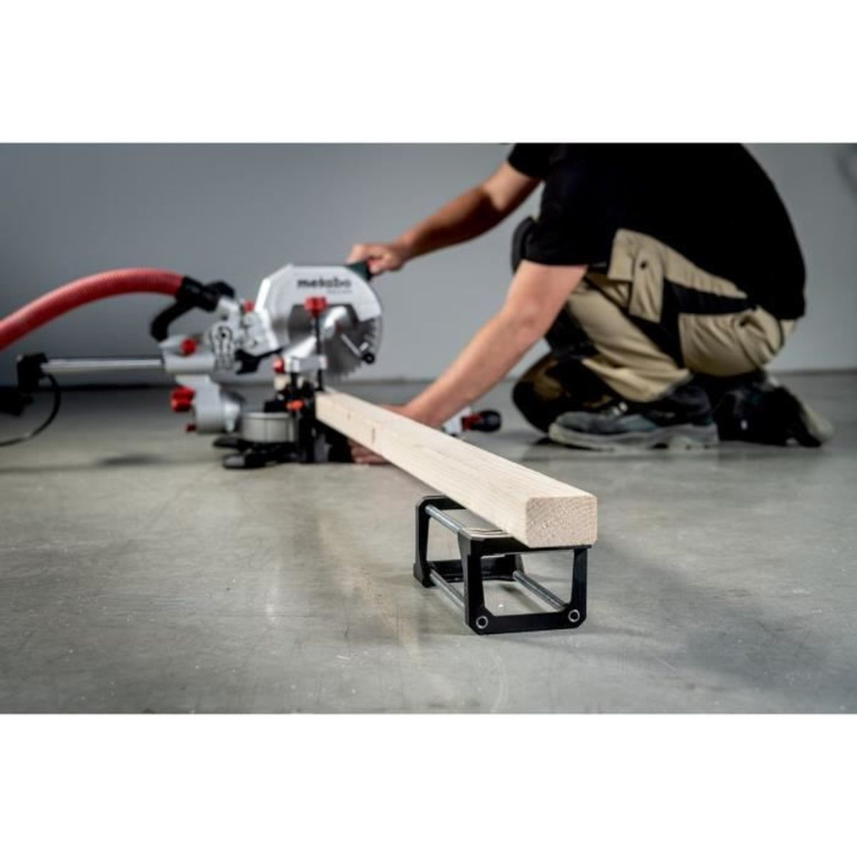 Een persoon gebruikt een zaag om een houten plank te zagen. De zaag is zilver en rood, en de persoon draagt zwart en kaki. Het werk wordt op beton gedaan.