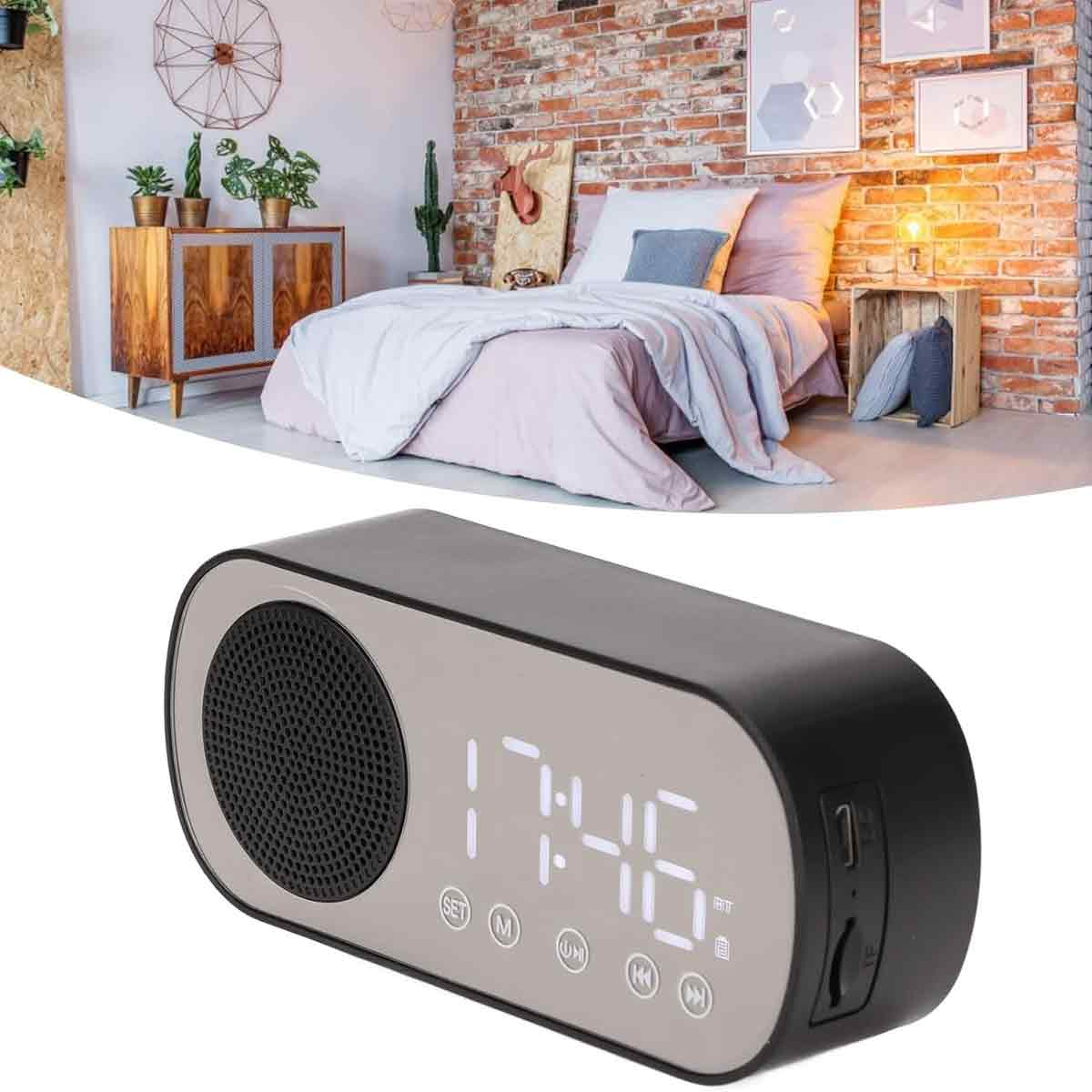 Eine schwarz-graue Digitaluhr mit Lautsprecher vor einem Schlafzimmer mit Bett und Backsteinmauer.