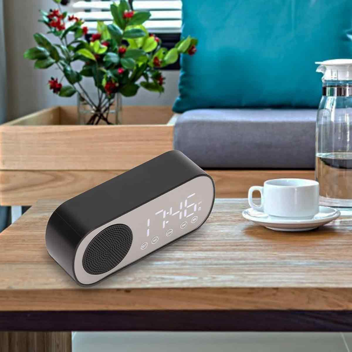 Ein schwarzer Lautsprecher mit Digitaluhr steht auf einem Holztisch. Eine weiße Tasse, Pflanzen und ein türkisfarbenes Kissen im Hintergrund.