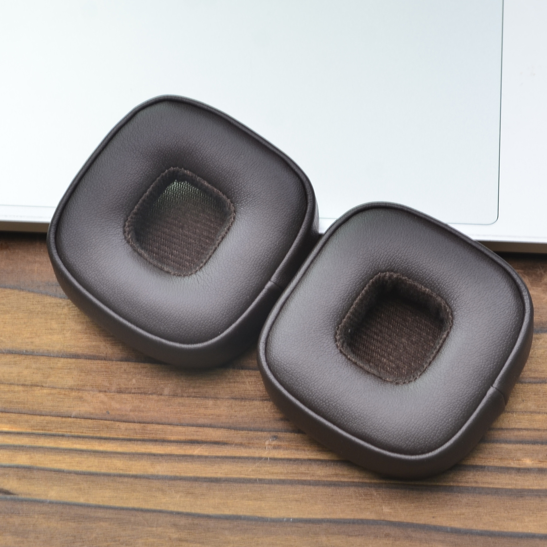 Zwei braune Leder-Kopfhörer-Ohrmuscheln auf einer Holzoberfläche neben einem Laptop. Jedes hat eine Stoffmitte.