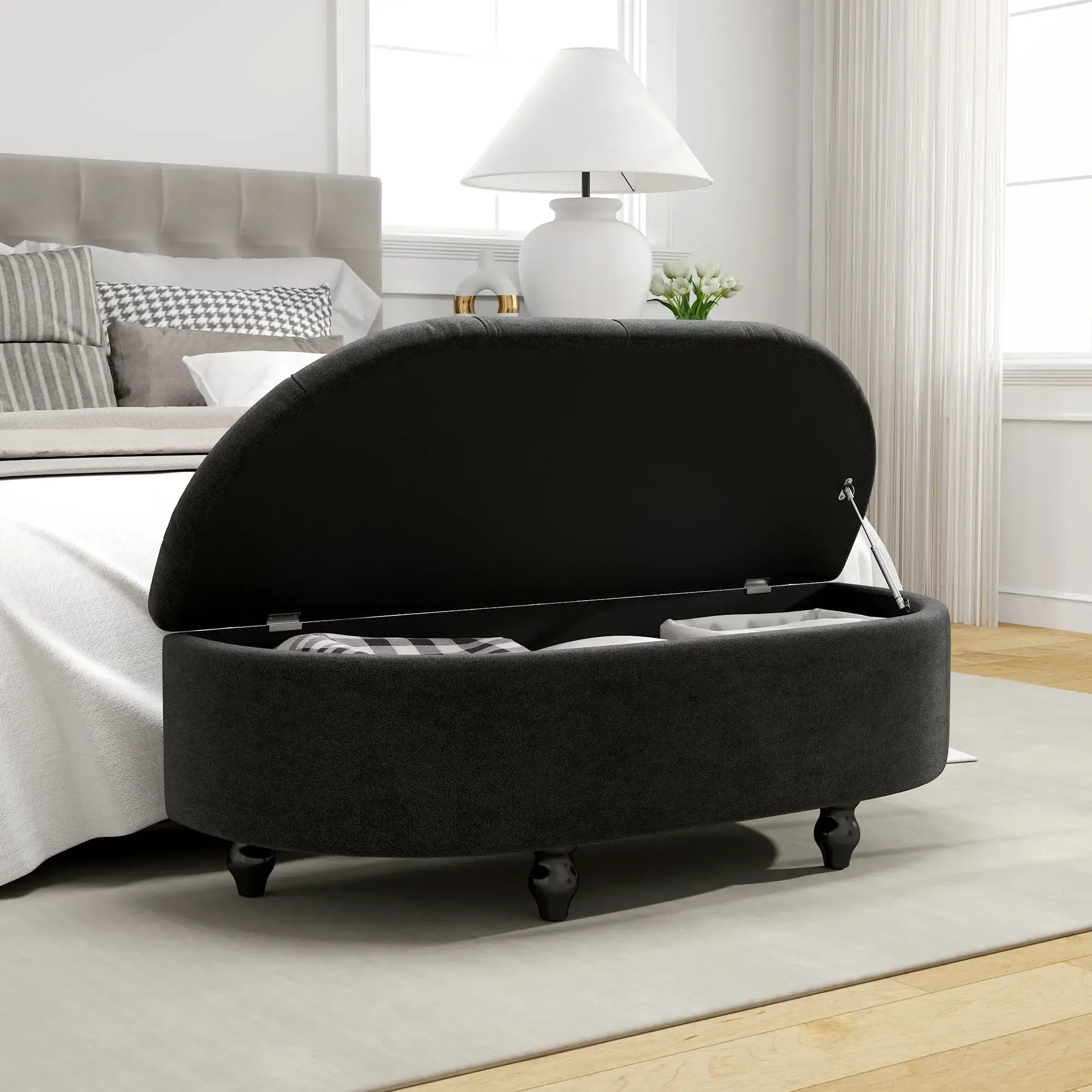 Eine schwarze Aufbewahrungshocker mit offenem Deckel steht in einem Schlafzimmer. Ein Bett, eine Lampe und ein Fenster sind im Hintergrund zu sehen.
