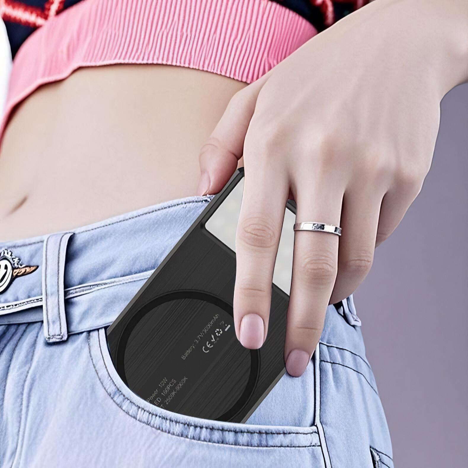 Eine Hand hält ein schwarzes Gerät in einer Jeans-Tasche. Die Person trägt einen Ring und ein rosa Top.