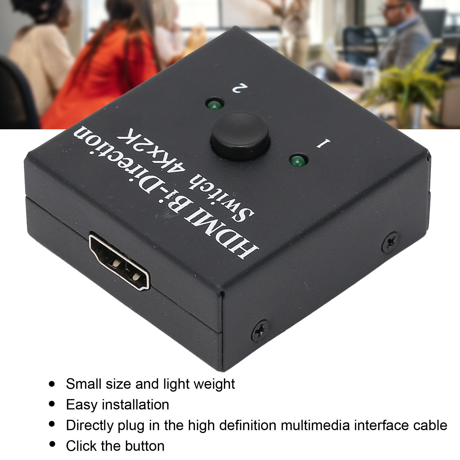 Ein schwarzer HDMI-Switch mit Knopf, zwei Kontrollleuchten und einem HDMI-Anschluss. Verschwommener Hintergrund mit Personen.