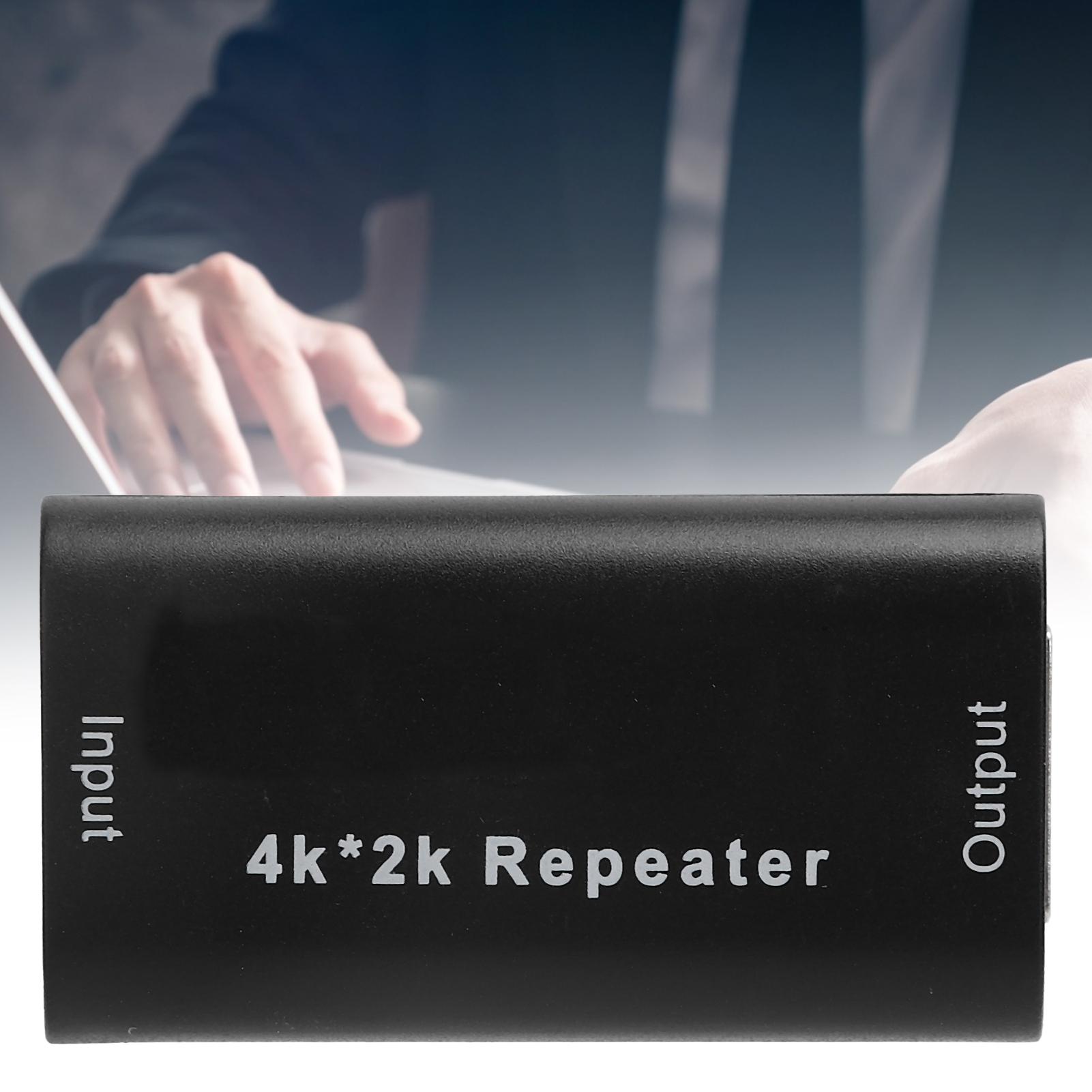 Ein schwarzer 4K*2k Repeater mit den Bezeichnungen Input und Output. Im Hintergrund ist verschwommen eine Person mit Laptop.