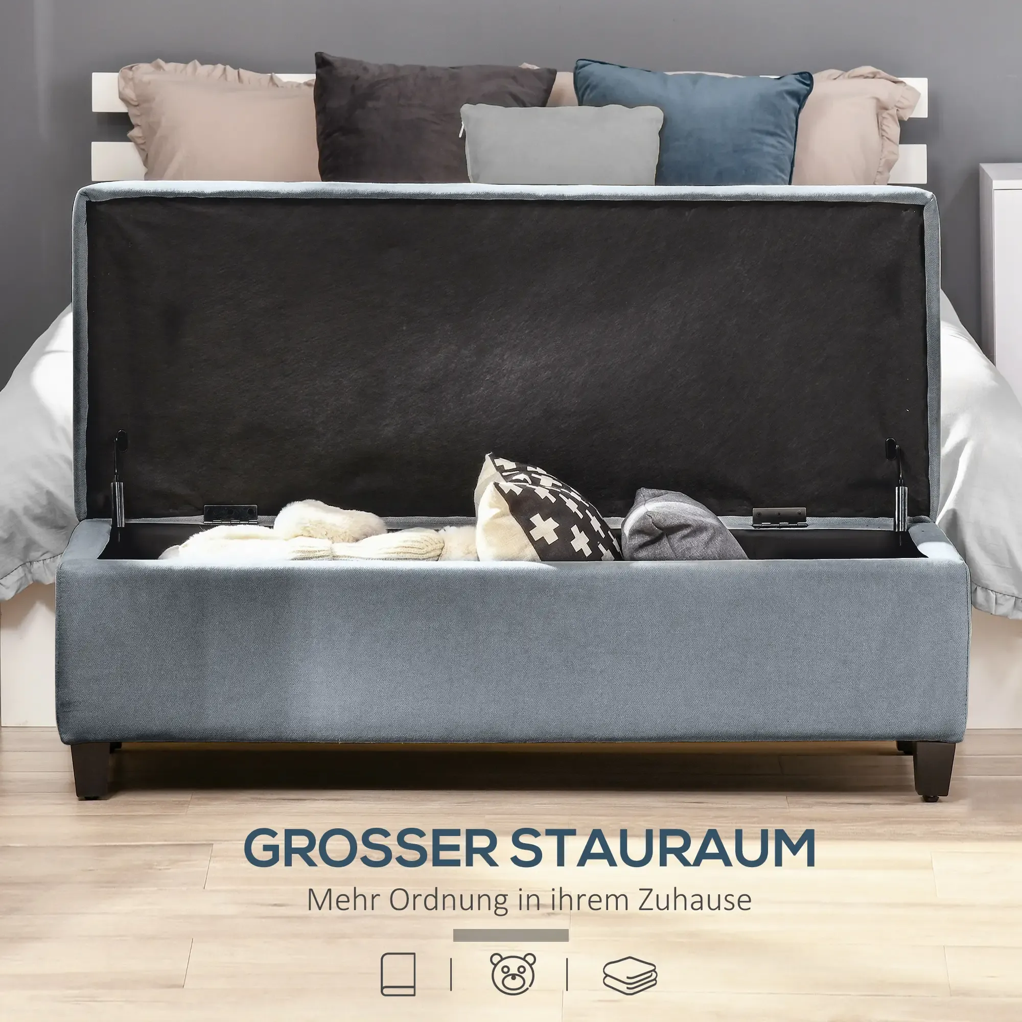 Eine blaue Stauraum-Ottomane mit offenem Deckel, die Decken und Kissen zeigt. Kissen befinden sich im Hintergrund auf einem Bett.