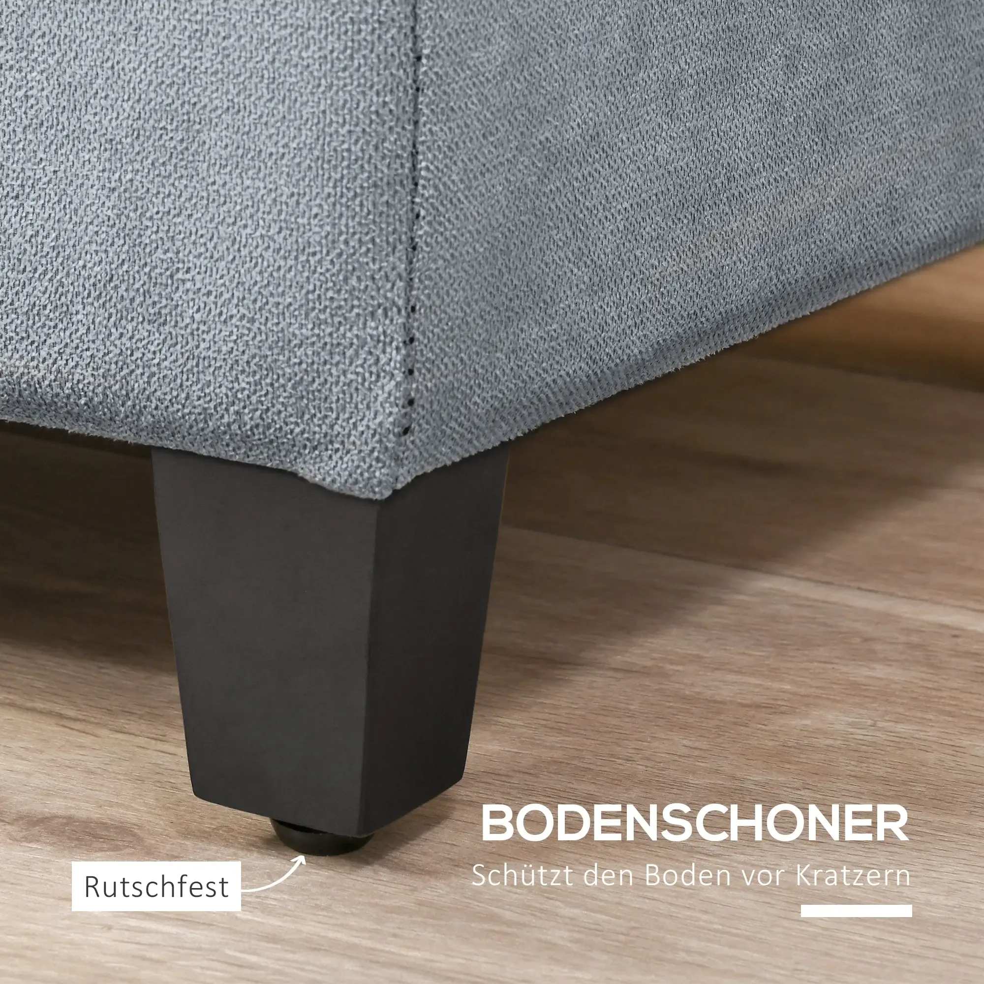 Nahaufnahme einer grauen Stoffsofa-Ecke mit einem schwarzen Bein. Das Bein hat einen Gummifuß. Holzfußboden im Hintergrund.