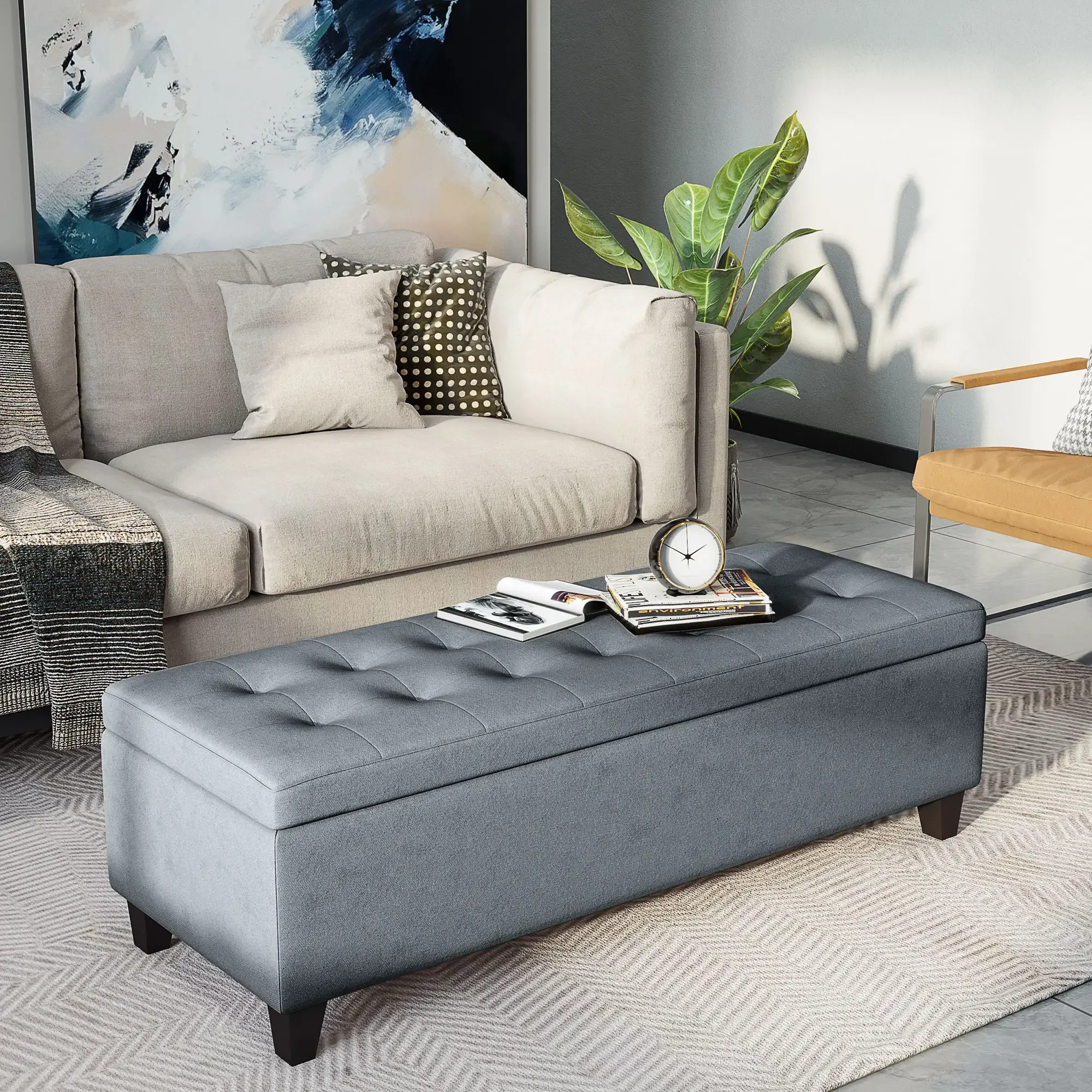 Eine graue Ottomane mit Büchern und einer Uhr steht vor einem beigen Sofa. Über dem Sofa hängt ein Gemälde.