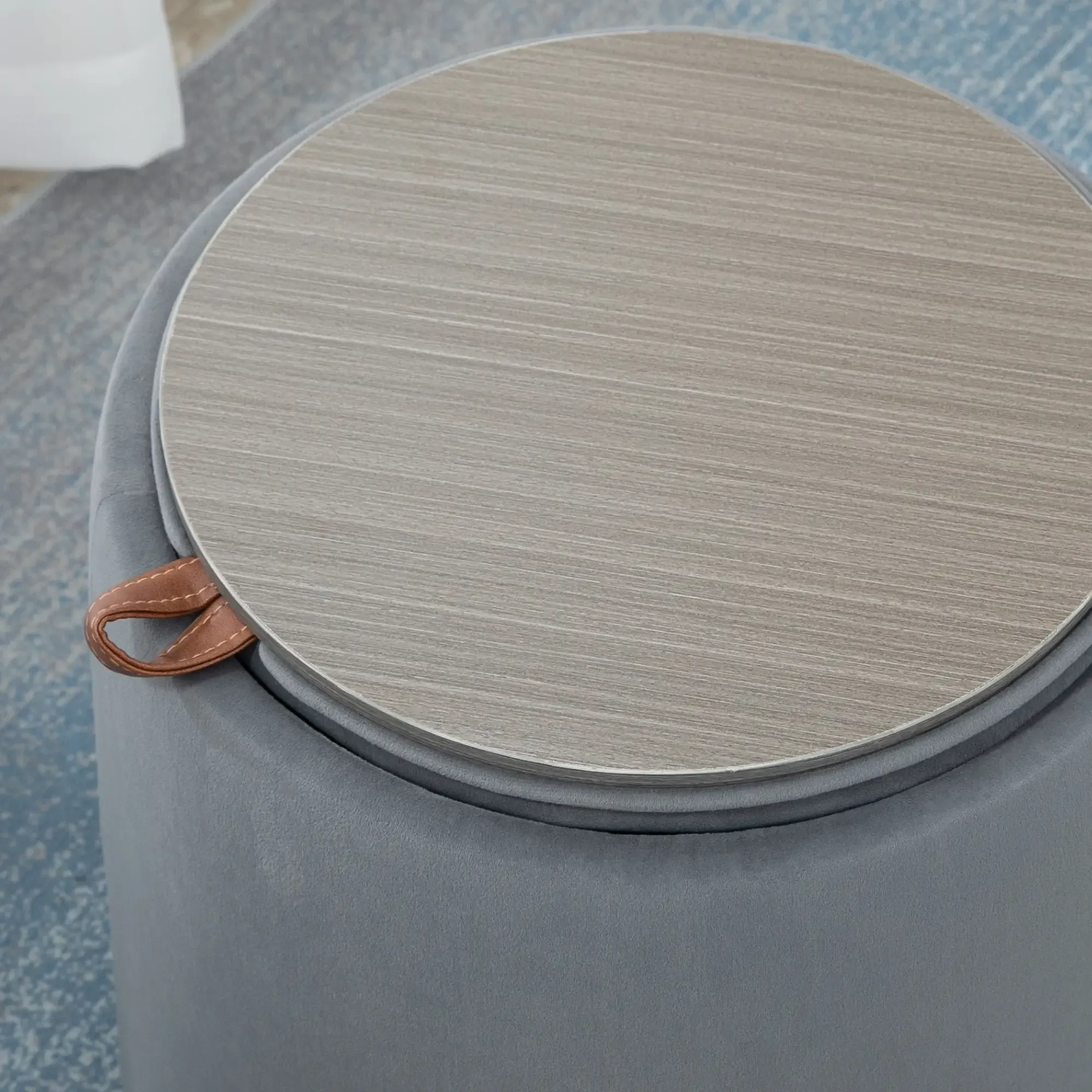 Ein runder, grauer Hocker mit heller Holzplatte. Ein brauner Ledergriff ist an der Seite befestigt. Der Hocker steht auf einem blauen Teppich.